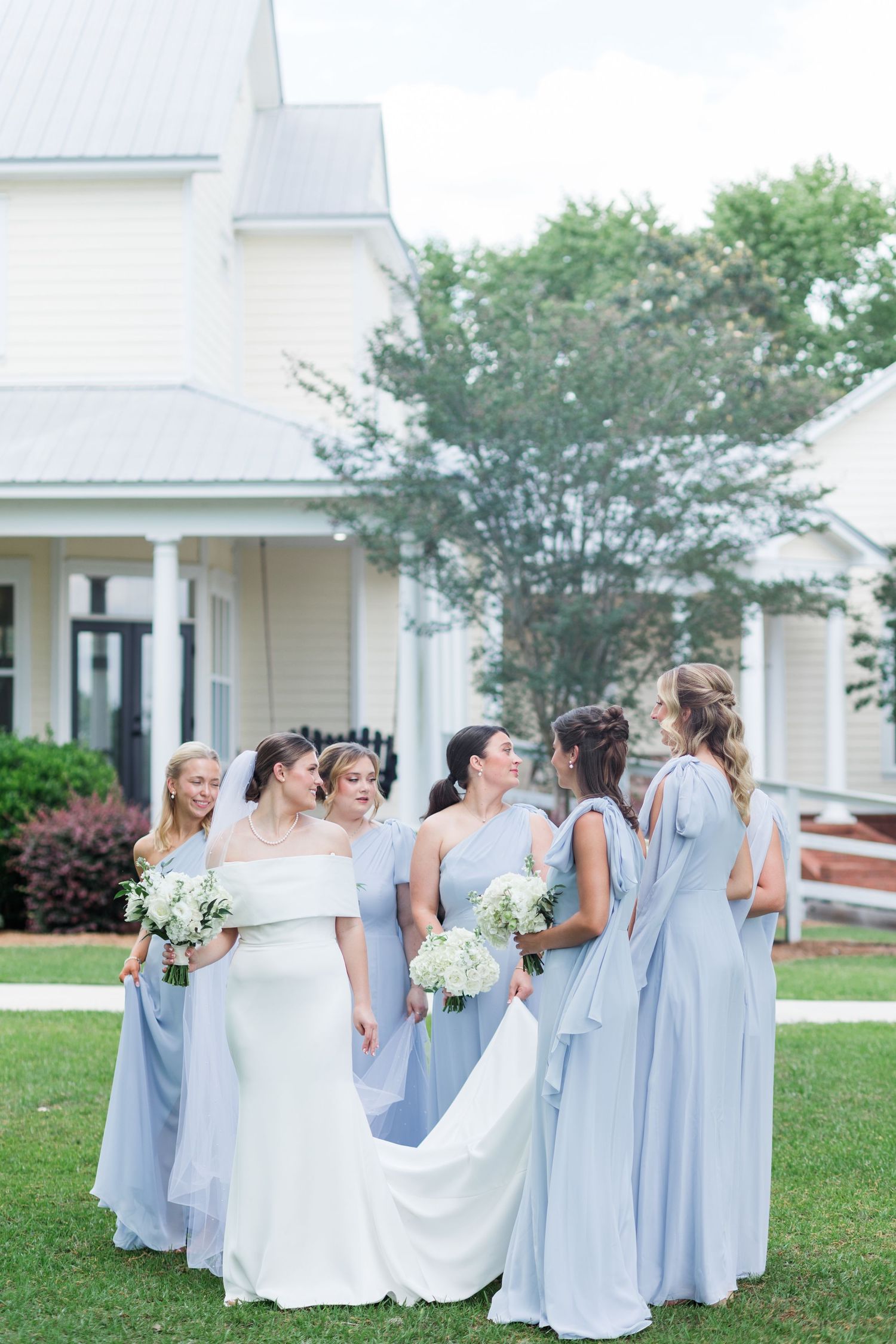 Bride surrounded by bridesmaids in periwinkle blue gowns holding white bouquets outside white chapel.