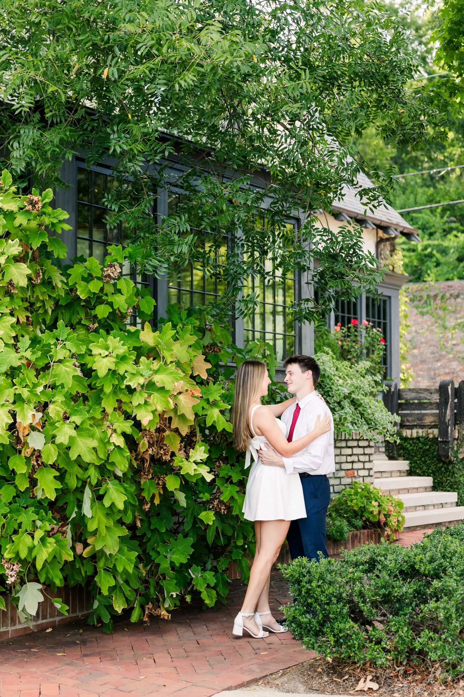 Couple embracing in lush garden setting beside ivy-covered building with brick pathway and green foliage.
