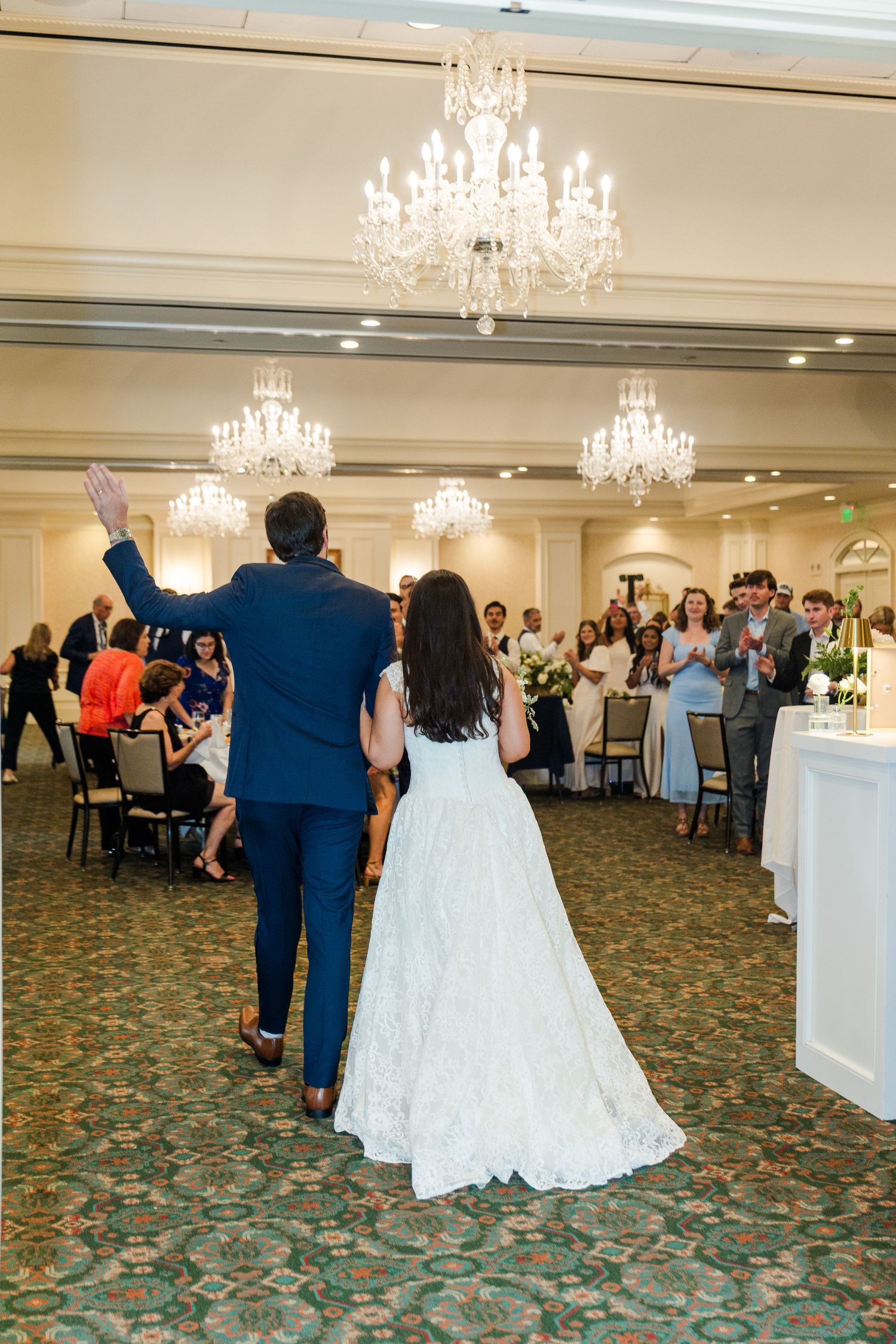 Bride and groom making grand entrance into elegant ballroom with crystal chandeliers and applauding guests.