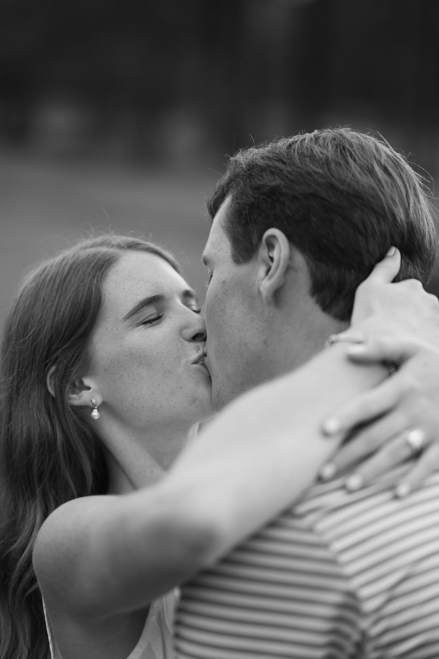 Black and white photo of a couple sharing a romantic kiss, woman with earrings embracing man in striped shirt outdoors.