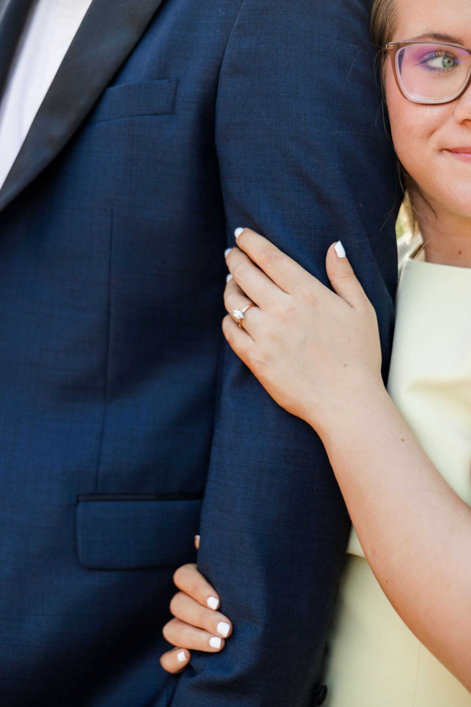Woman in glasses wearing engagement ring clings to mans navy suit jacket sleeve during engagement photo session.