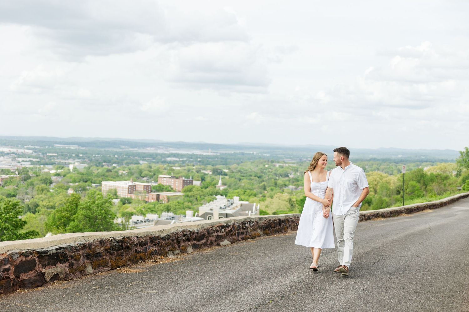 Couple walks hand-in-hand on scenic overlook road with panoramic city and green landscape views under cloudy sky.