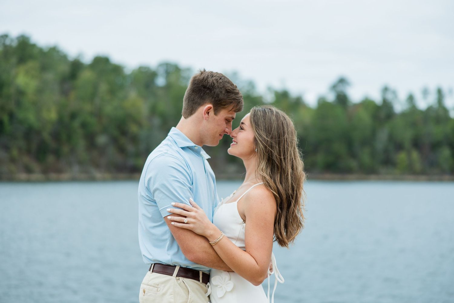 Engaged couple standing nose to nose in a romantic embrace beside a lake with a forested shoreline in the background.