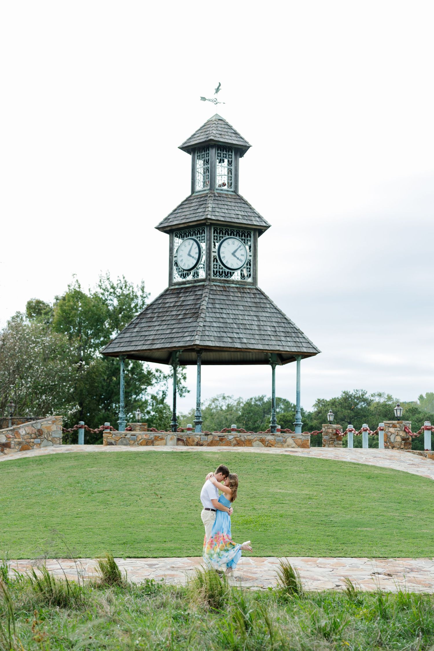 Engaged couple embraces on a stone path with a grassy hill and a historic clock tower gazebo in the background.