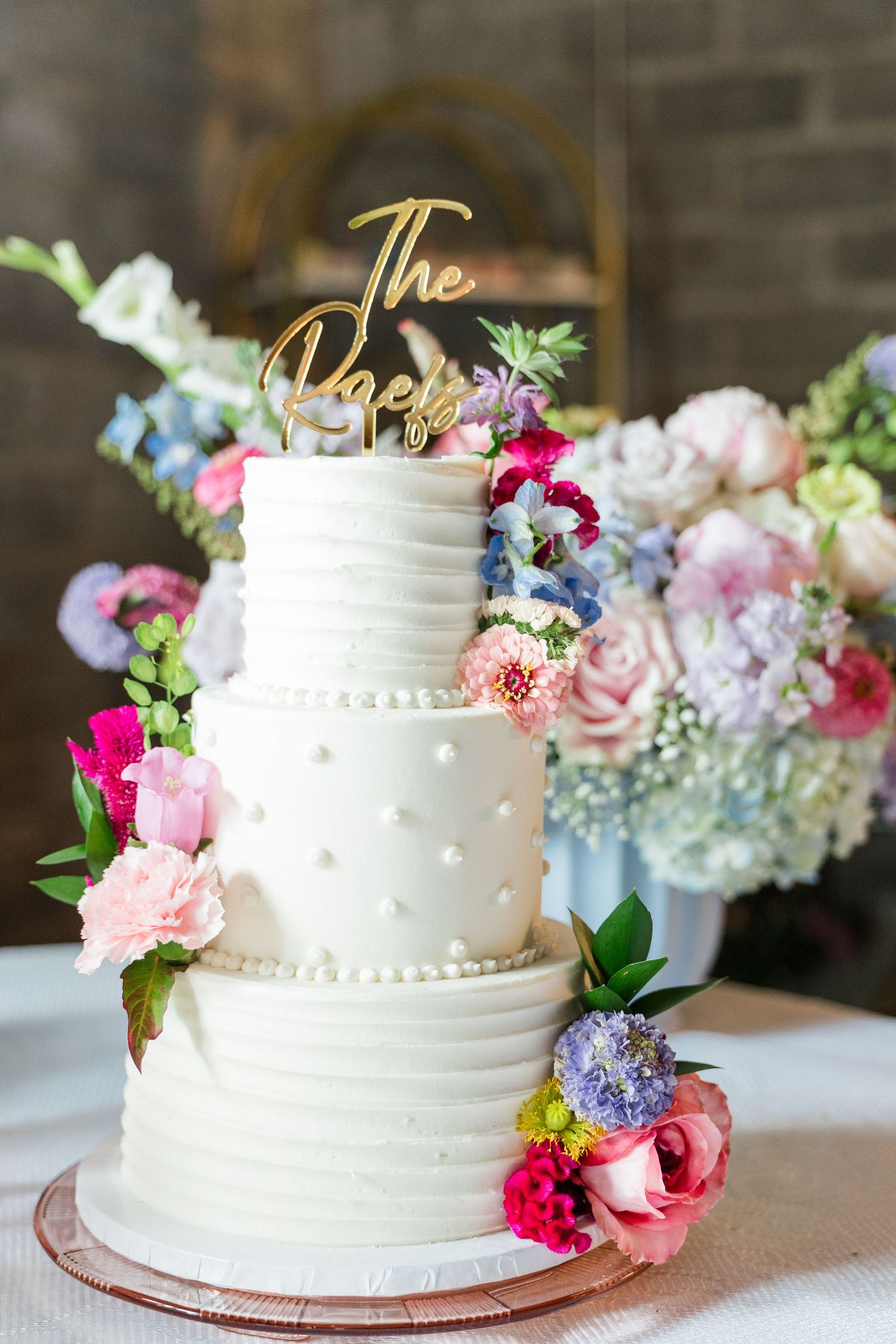 Three-tier white wedding cake with pearl details, floral decorations, and gold cursive cake topper surrounded by colorful blooms.