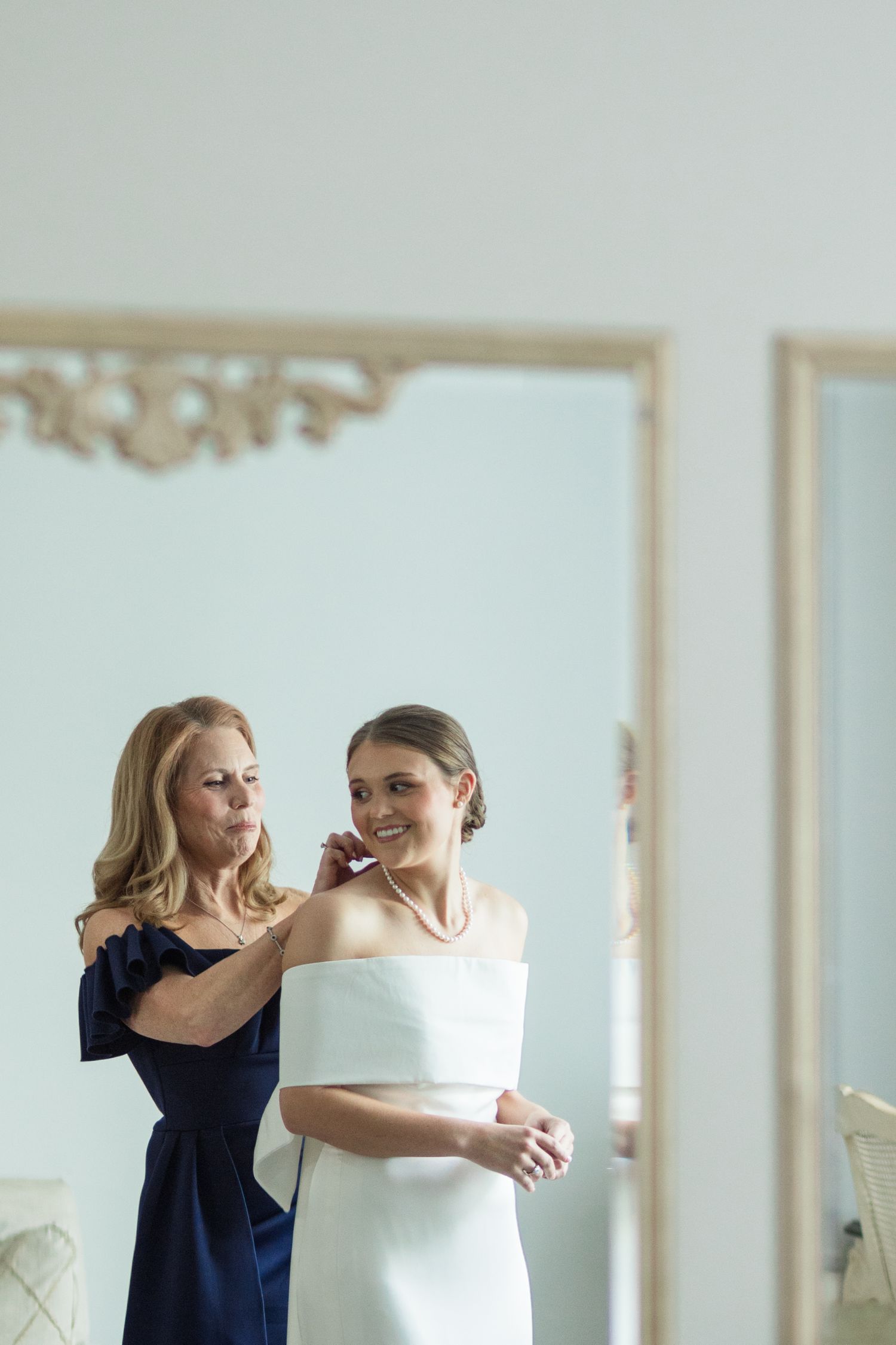A mother in navy blue helps her smiling bride daughter in a white off-shoulder gown with jewelry before the wedding.