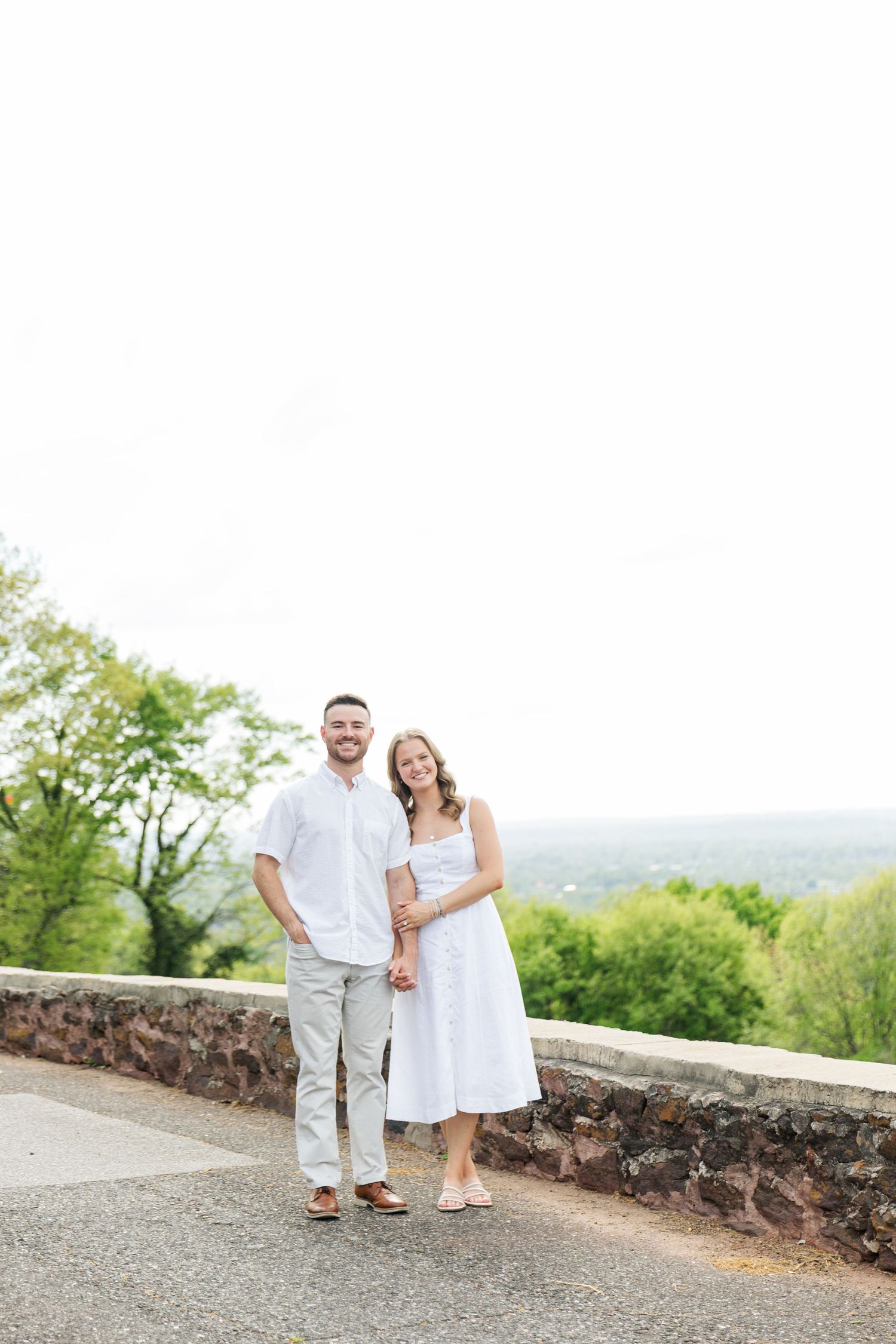Couple in matching white outfits holding hands on a scenic overlook with lush green trees in the background.