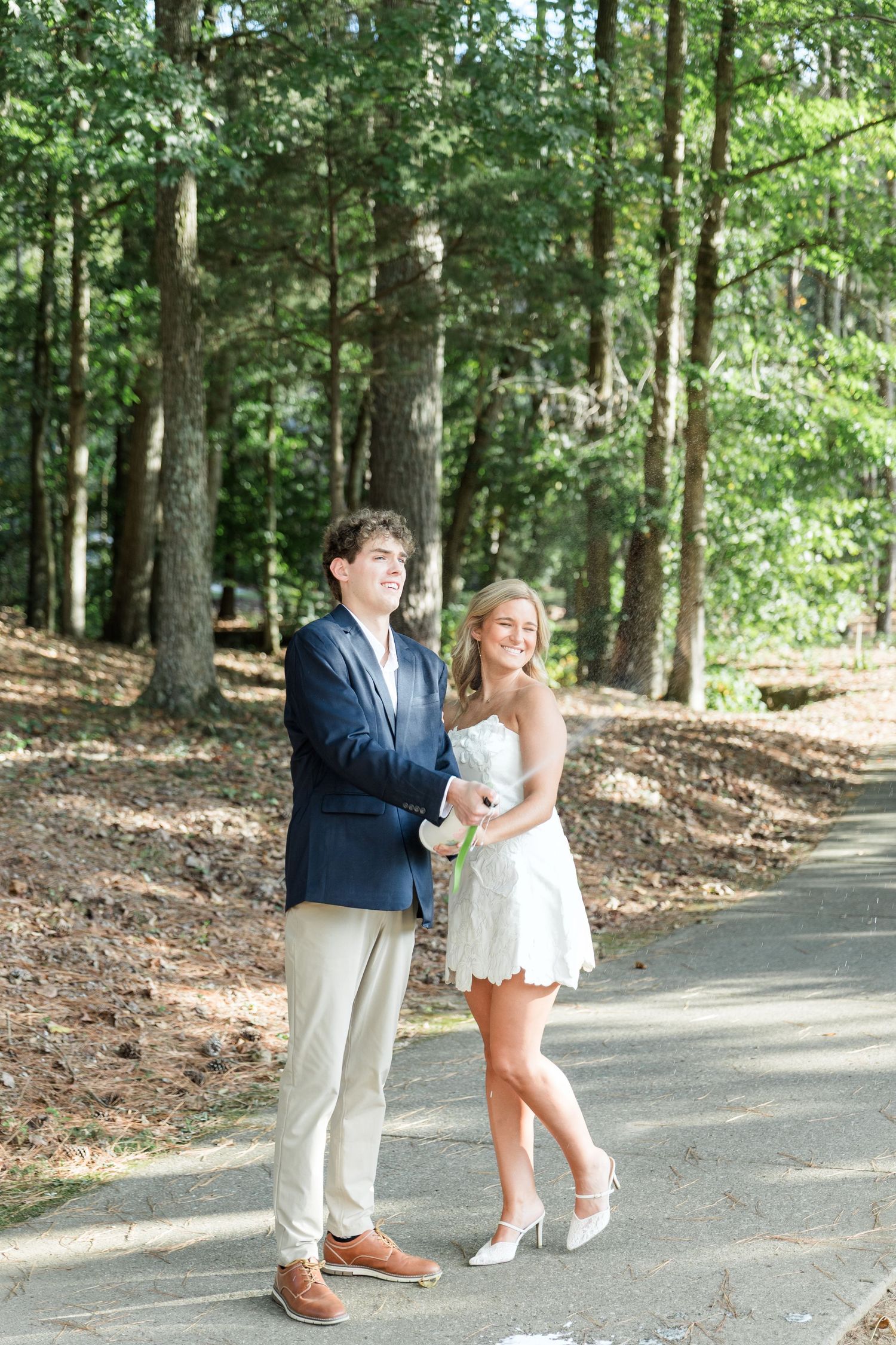 Happy engaged couple holding flowers and posing together on a tree-lined path, man in navy blazer and woman in white dress.