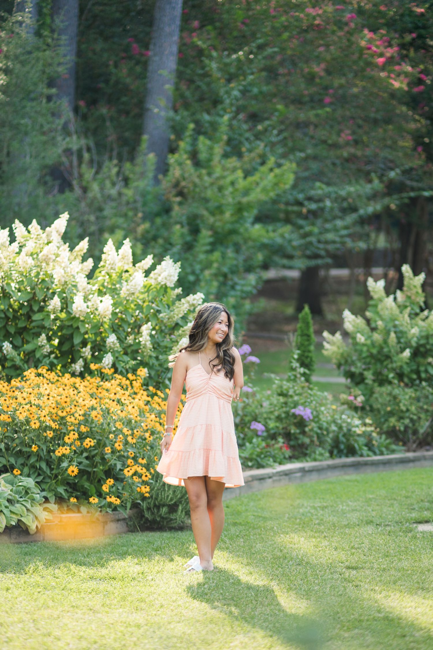 Young woman in pink dress walking through lush garden with yellow flowers and white blooms.