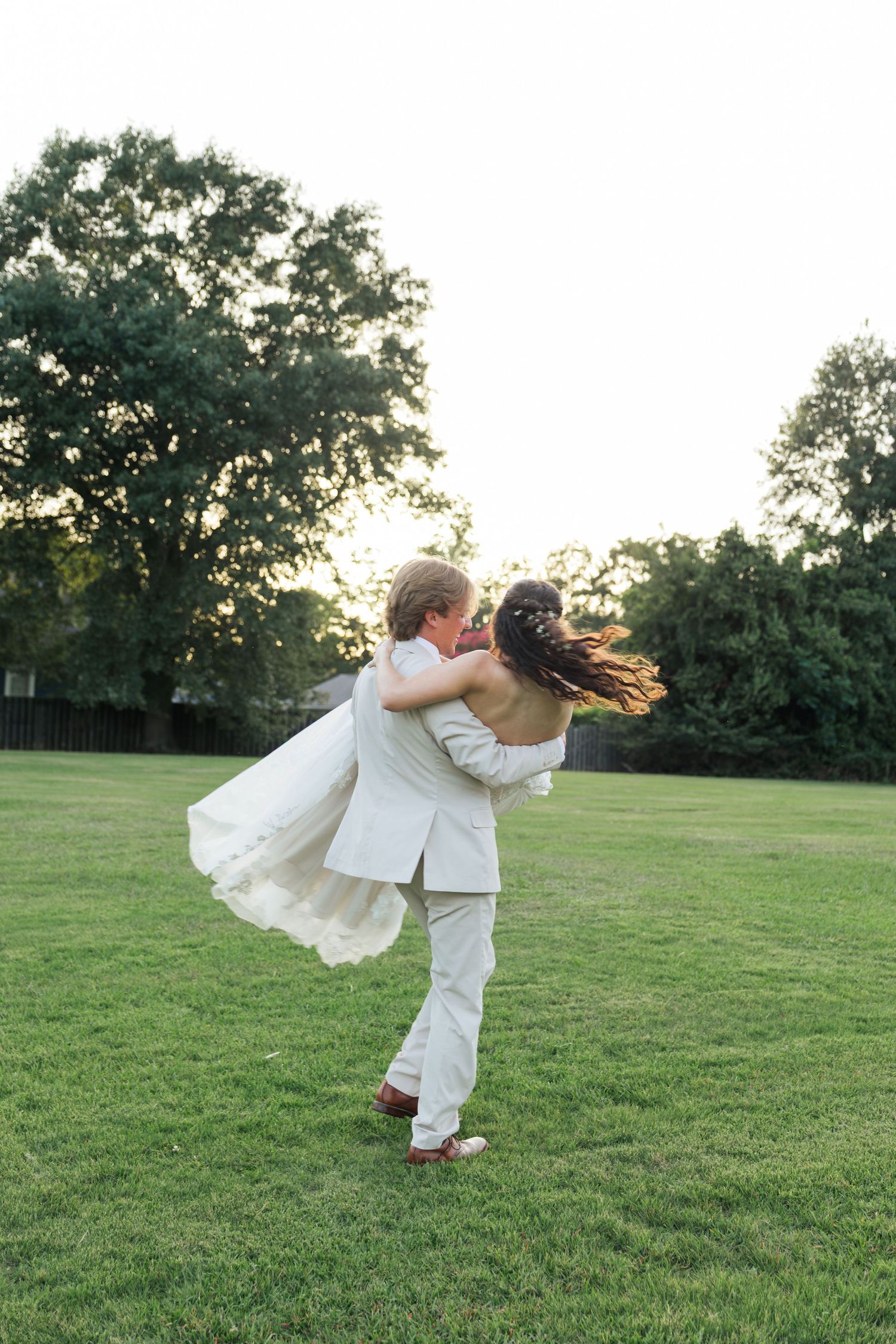 Groom lifting bride in white wedding dress at sunset in lush green outdoor field.