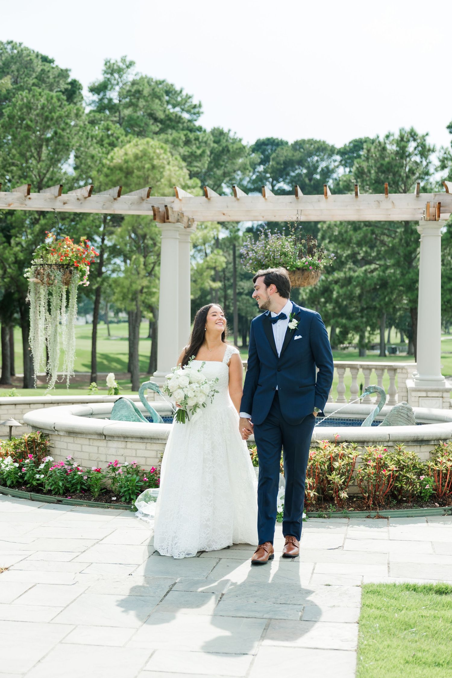 Bride and groom hold hands near a fountain under a pergola, surrounded by lush greenery on their wedding day.