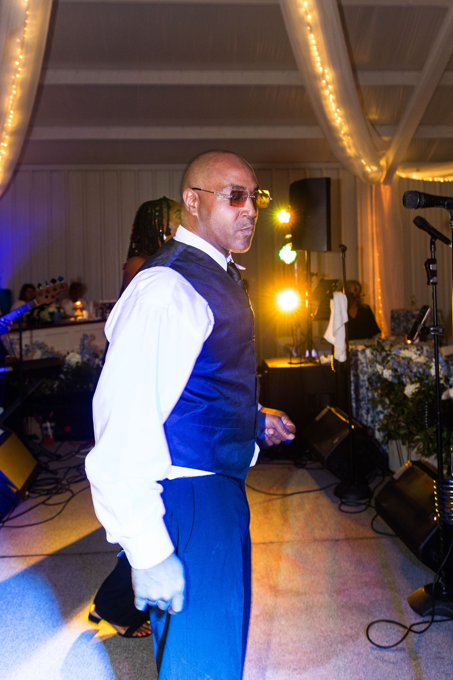 Energetic guest in blue vest and sunglasses dances under warm string lights at lively wedding reception.