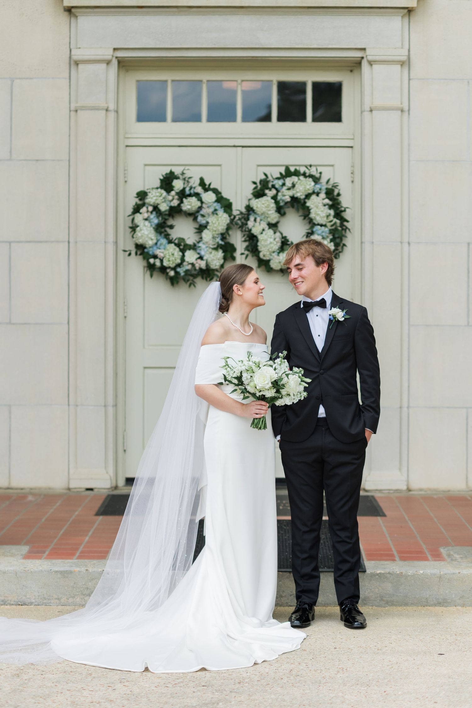 Bride and groom standing before white doors adorned with floral wreaths, bride holding white bouquet with flowing veil.