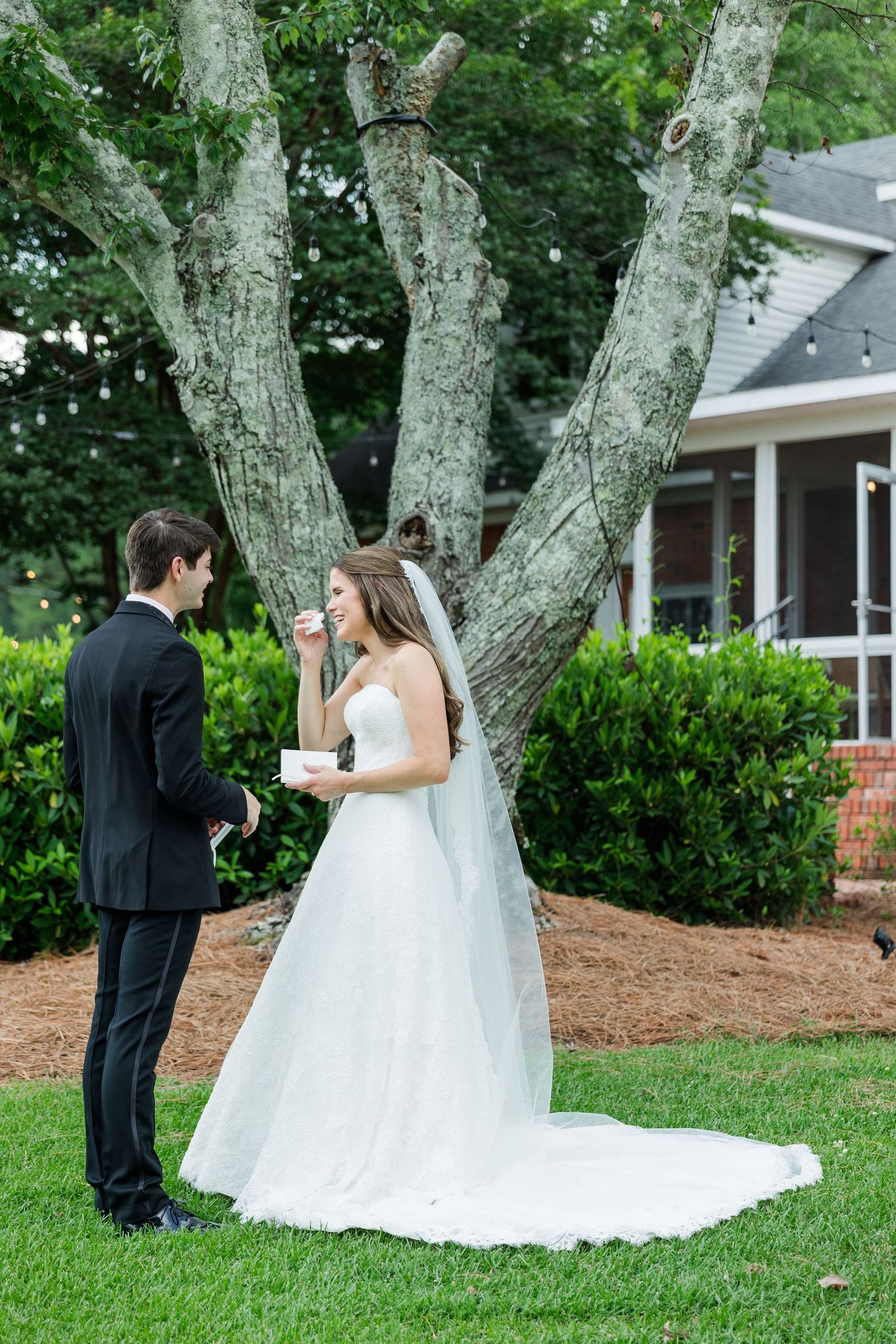 Bride in white gown reads vows while groom in black tuxedo listens outdoors near a large tree.