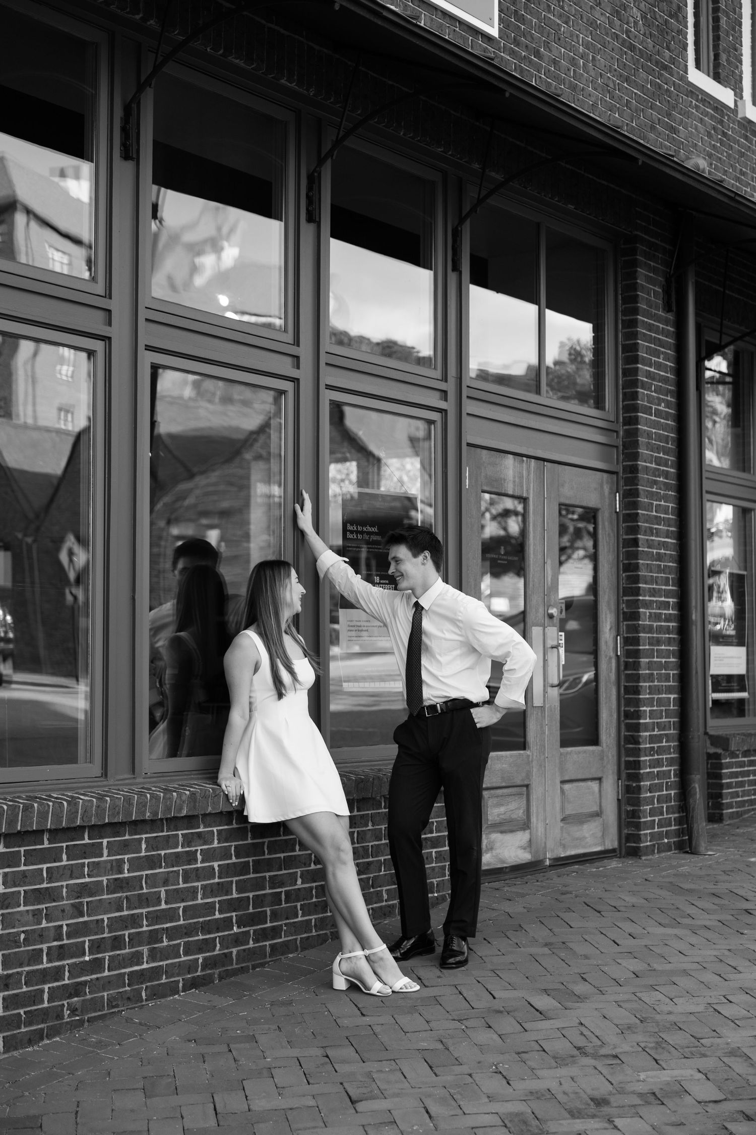 Black and white photo of a couple in formal attire sharing a romantic moment outside a brick building with large windows.