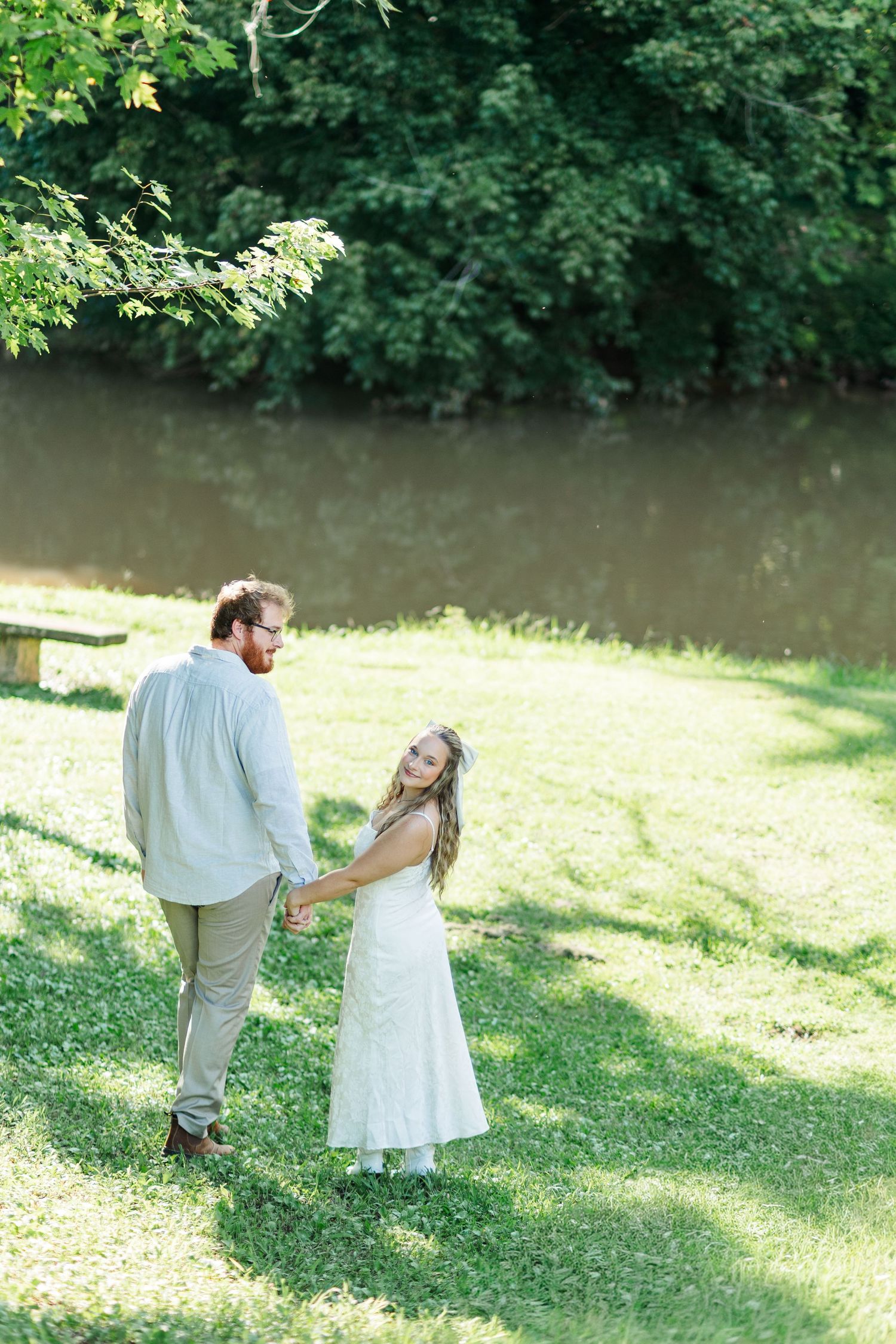 Couple holding hands walking by a serene river surrounded by lush green trees during an outdoor engagement session.