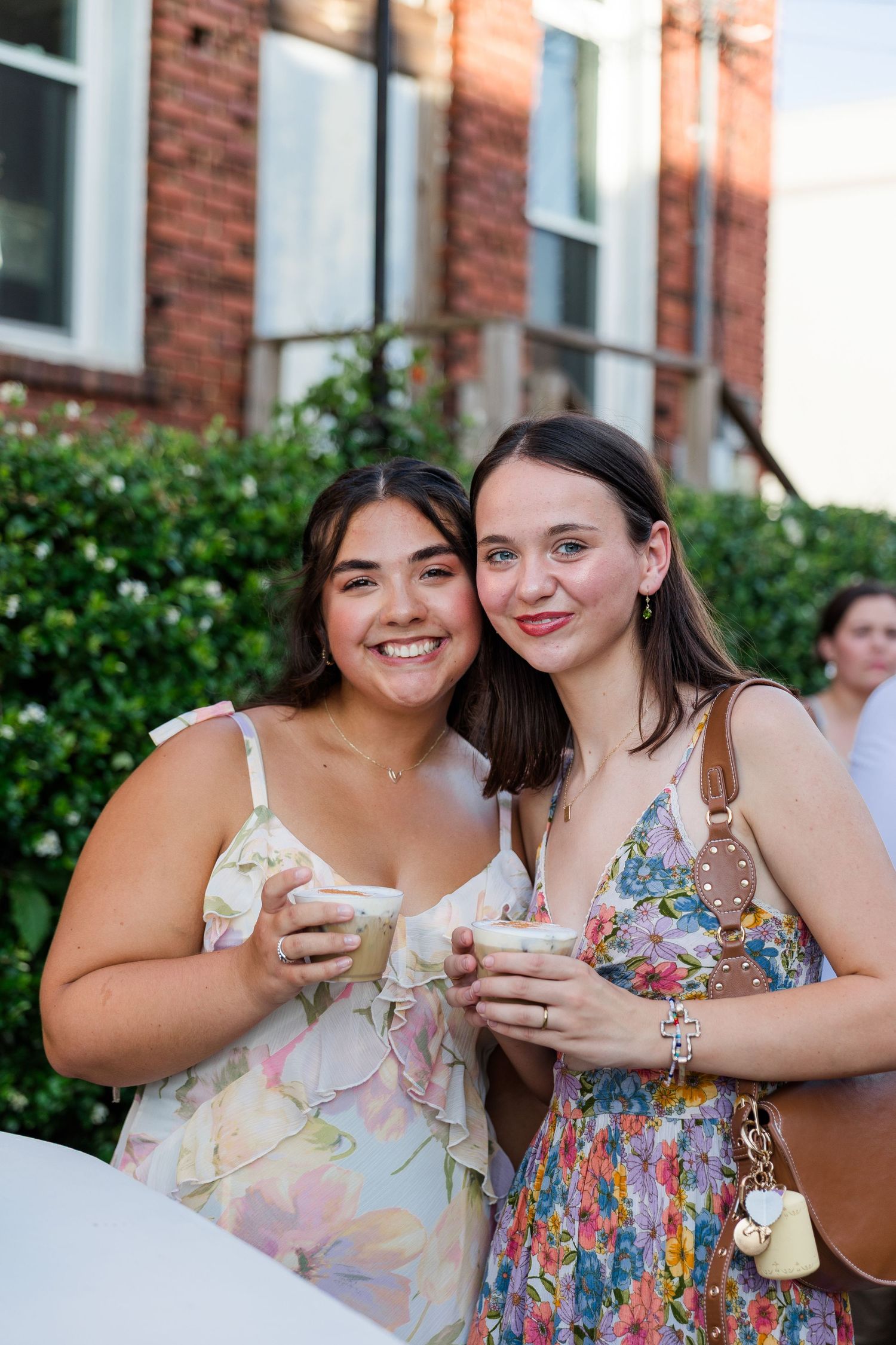 Two young women in floral dresses smiling and holding drinks at an outdoor social gathering.