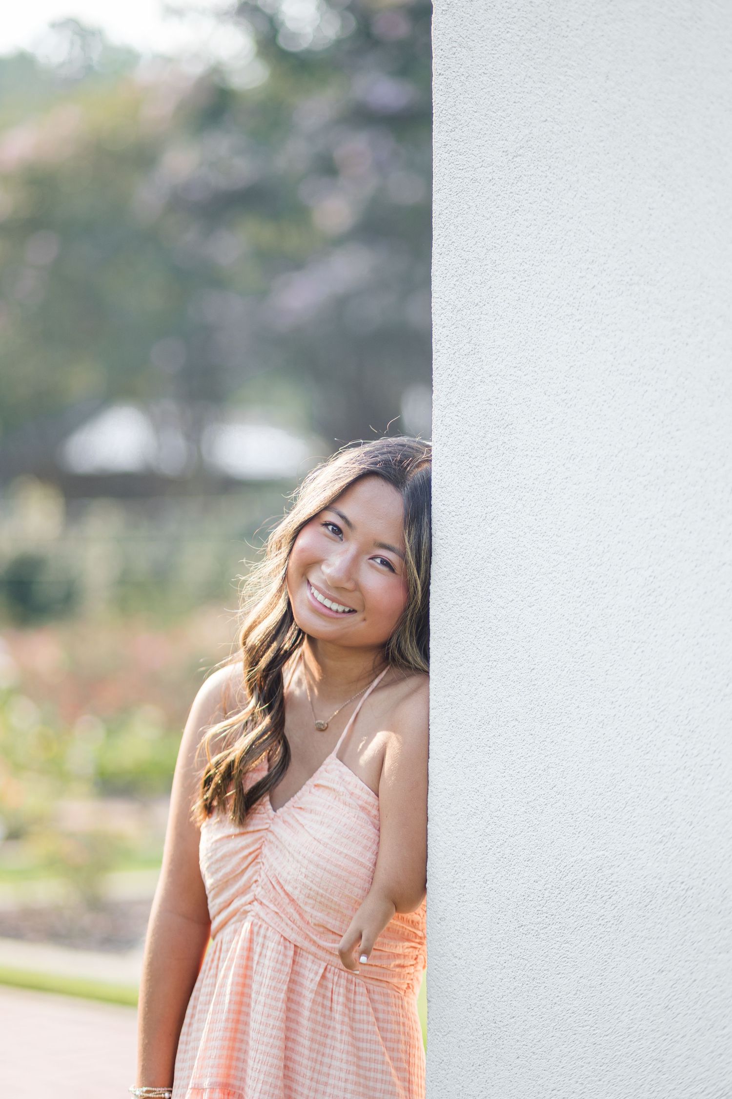 Smiling young woman in a peach sundress leaning against a white wall outdoors.