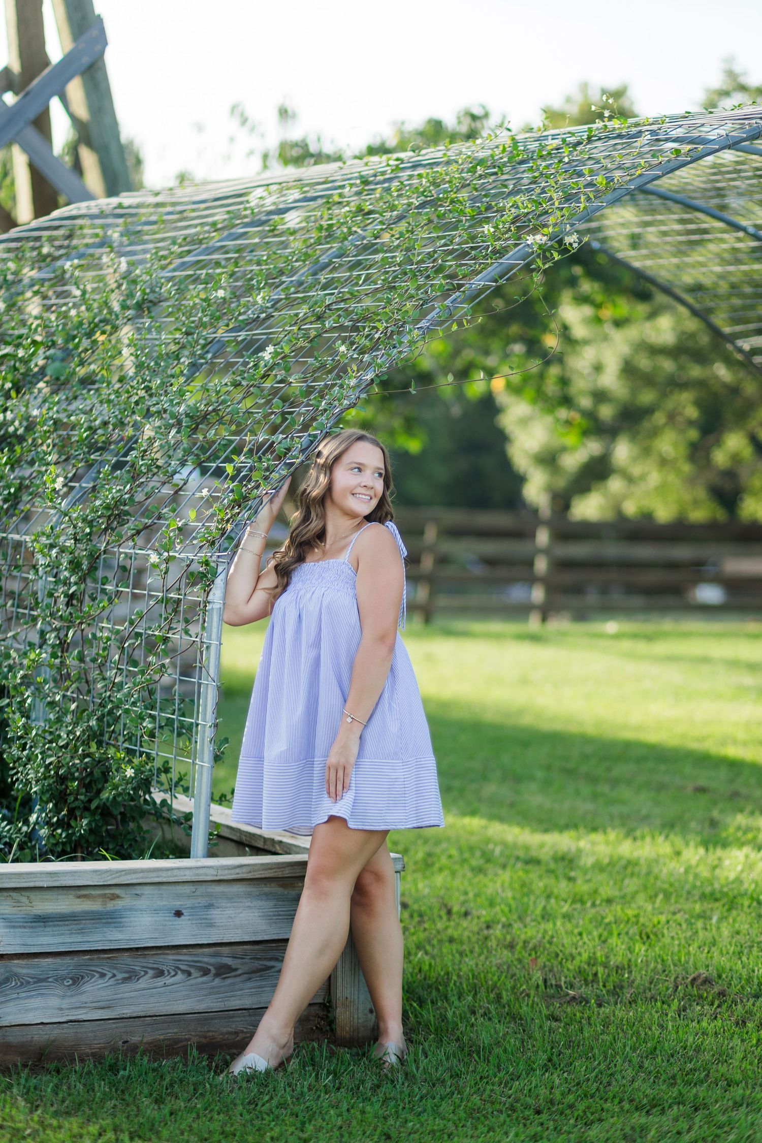 Young woman in light blue dress posing outdoors near a wire garden arch with lush greenery on a sunny day.
