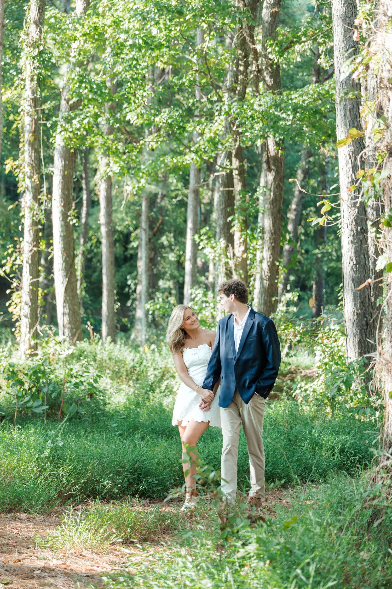 Couple holding hands and gazing at each other in a lush green forest during a summer engagement session.