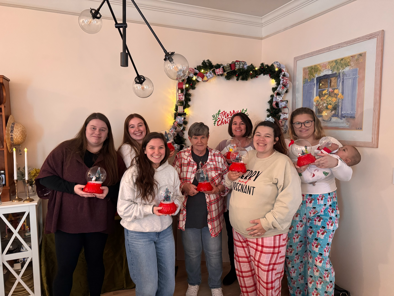 Group of women in holiday pajamas posing together in front of a Christmas decoration arch holding red gift boxes.