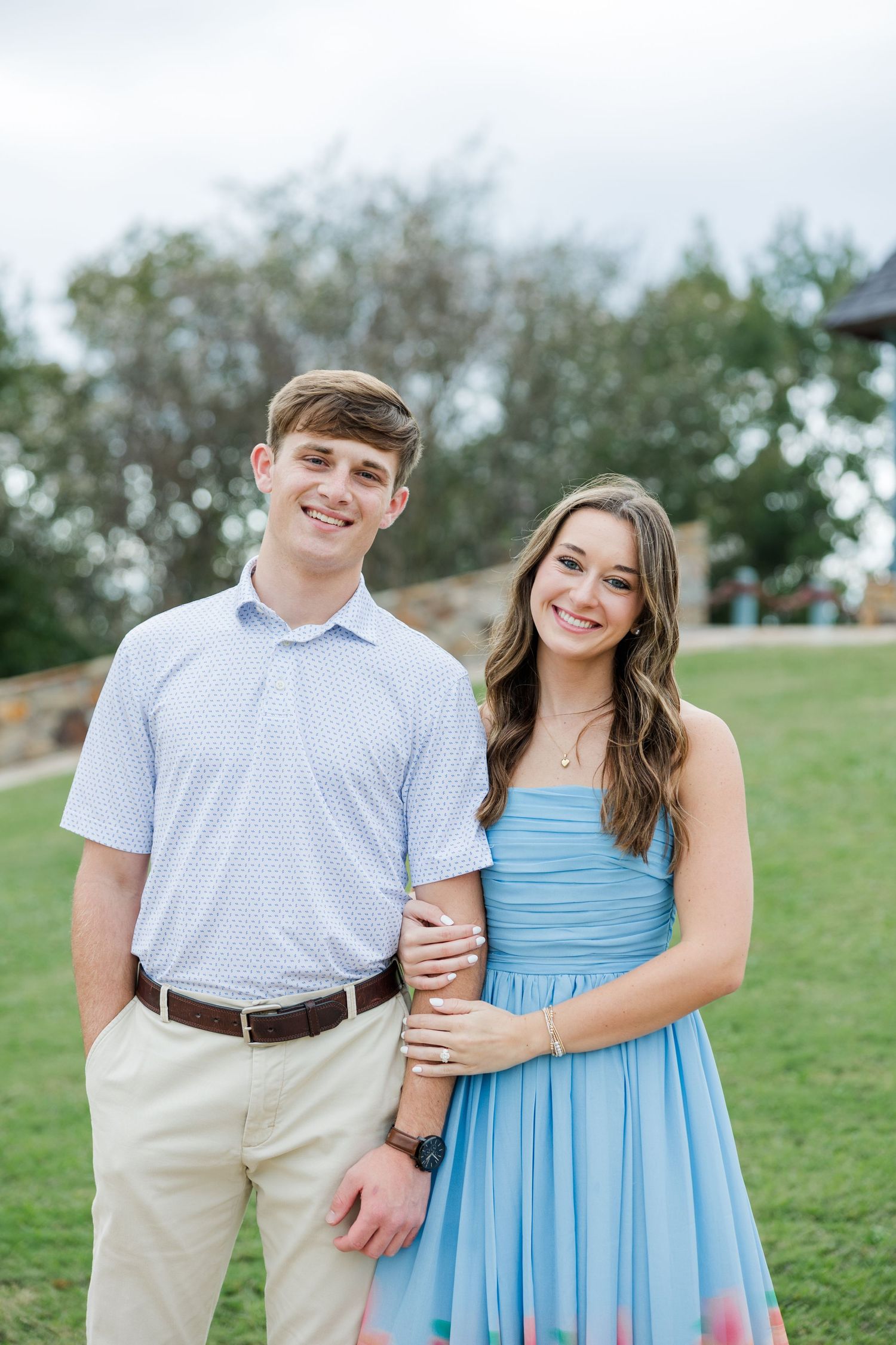 Smiling couple standing arm in arm on a green lawn, he in a light blue polo and khakis, she in a blue sleeveless dress.
