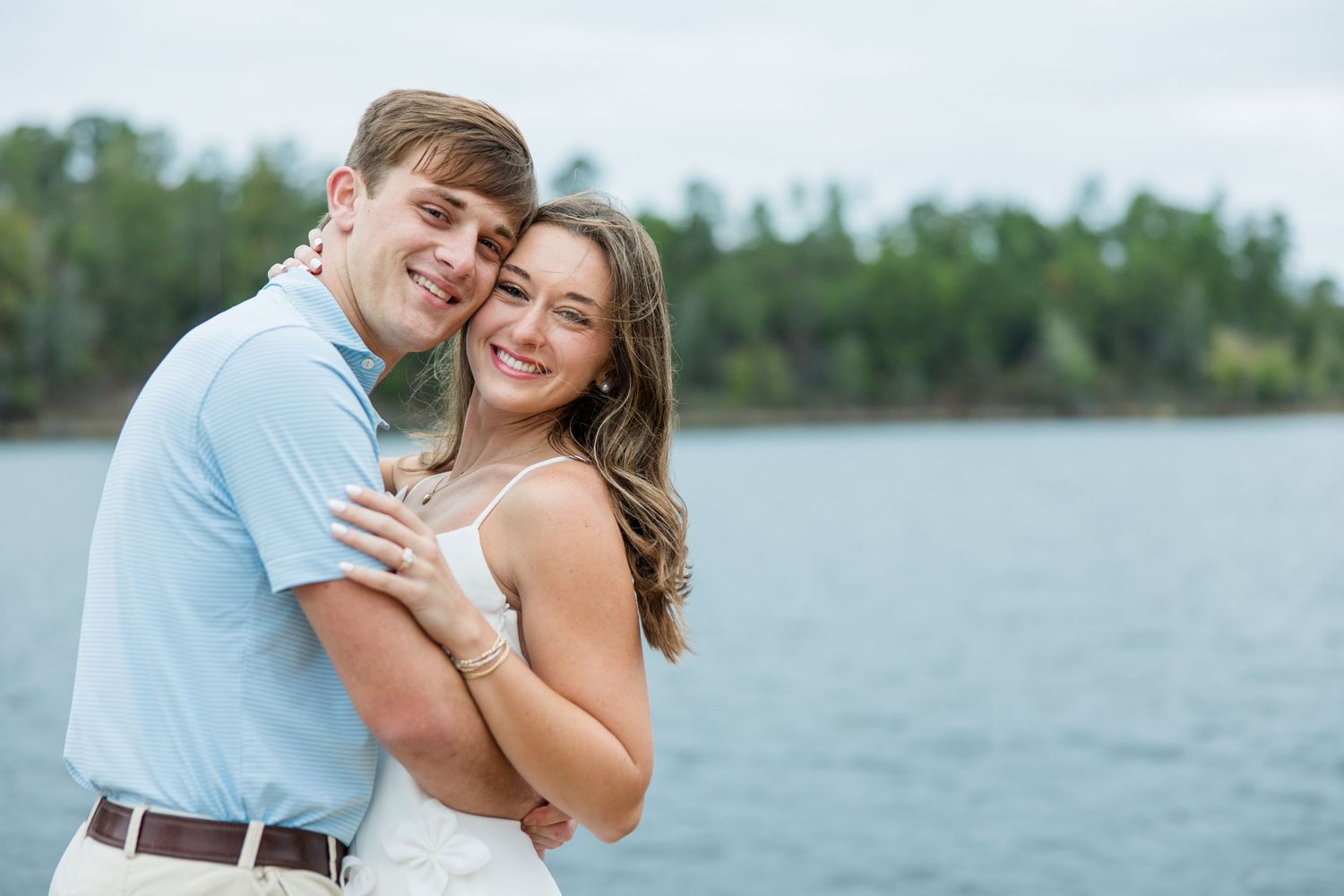 Smiling engaged couple hugging close in front of a calm lake and tree-lined shore during an outdoor engagement session.