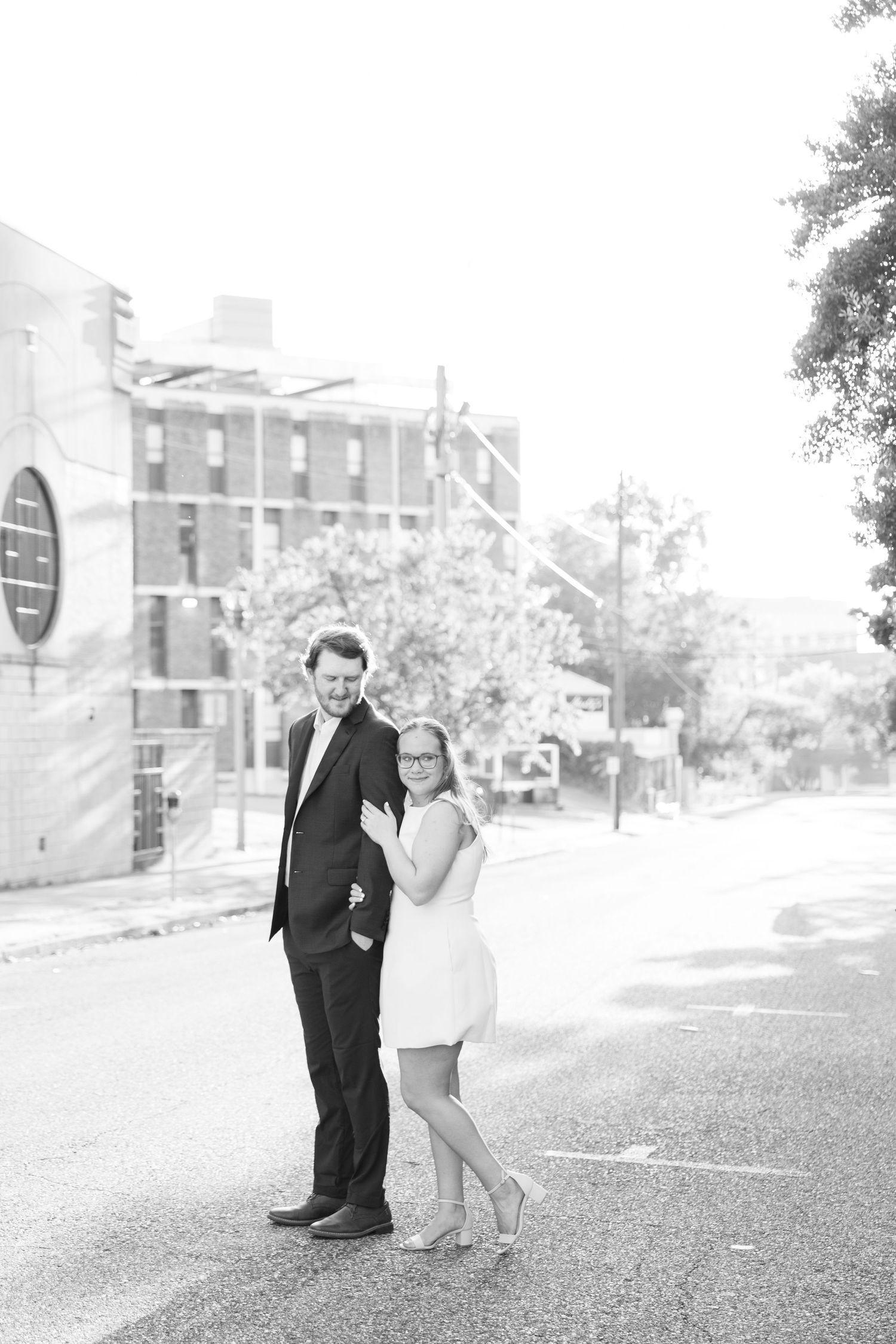 Black and white photo of a couple posing on a city street, woman in short dress embracing man in dark suit.