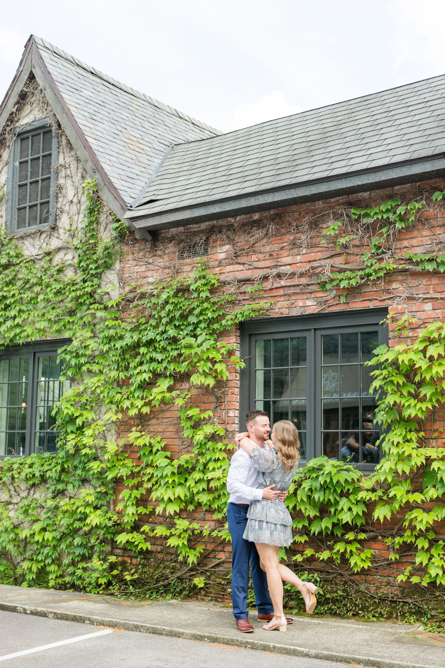 Couple embracing and dancing in front of ivy-covered brick building during outdoor engagement photo session.