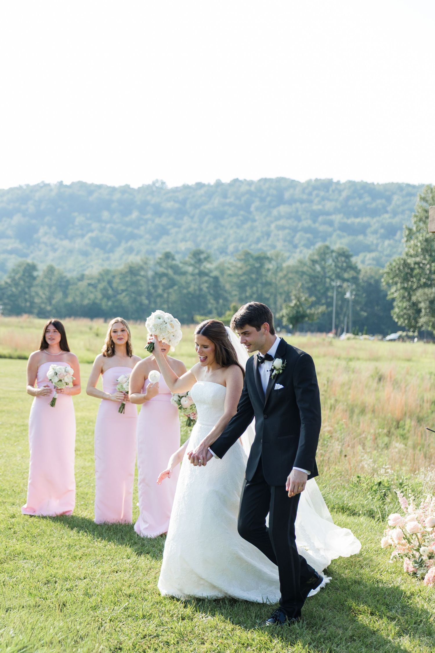 Bride and groom walk hand-in-hand on green lawn while three bridesmaids in pink gowns follow with bouquets.
