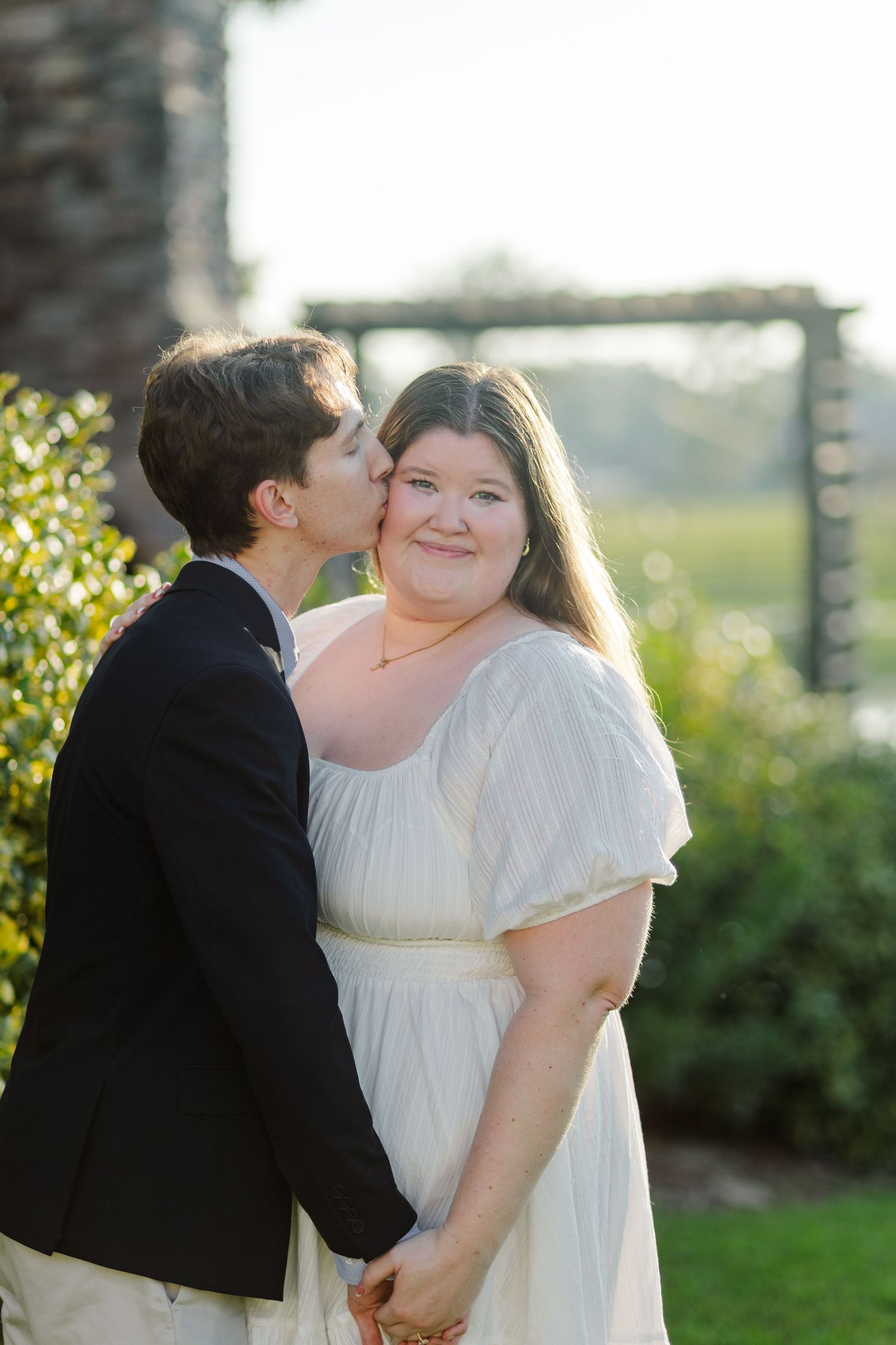 Groom in black suit kissing bride on cheek as she smiles in a white dress outdoors.
