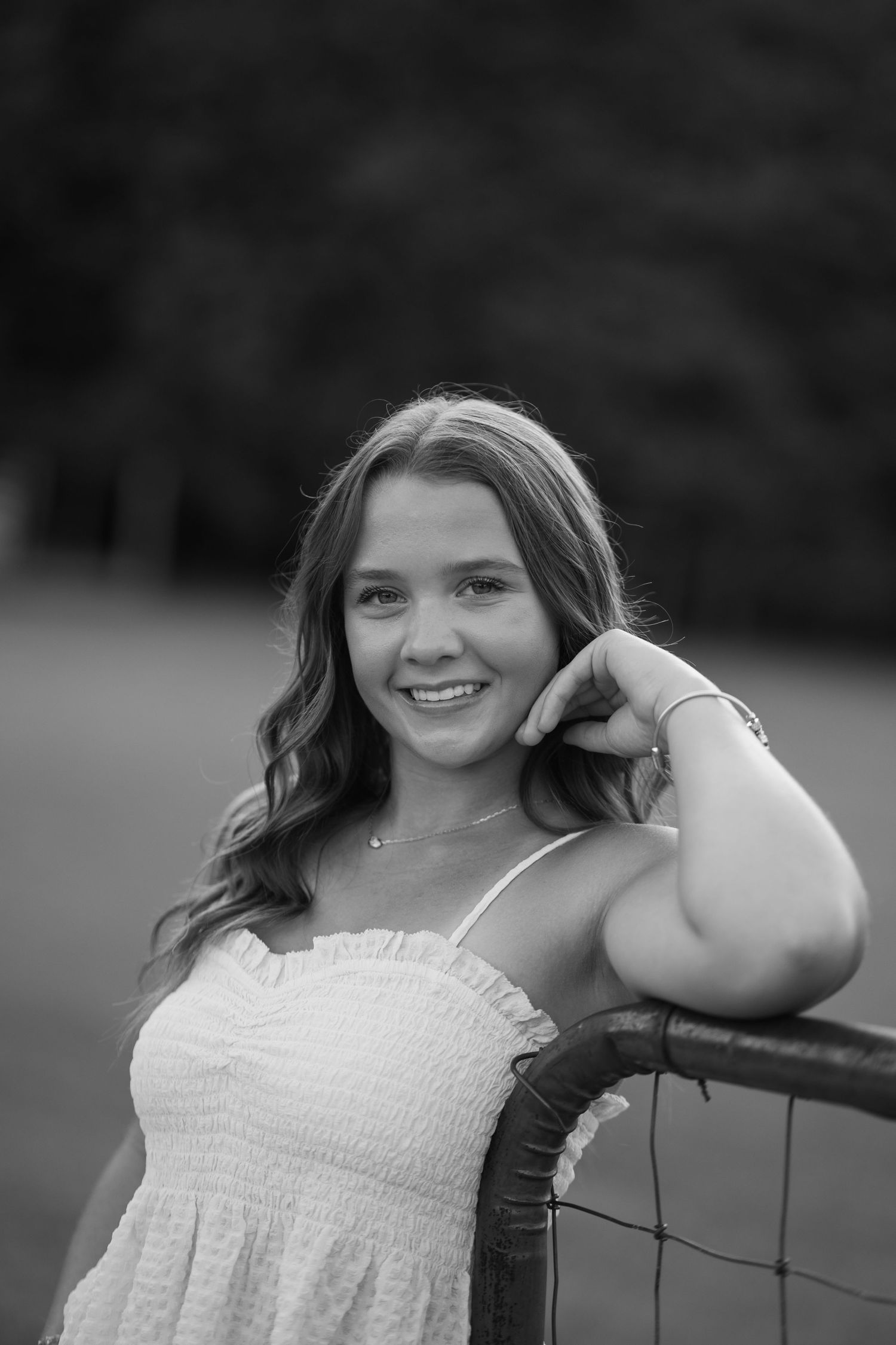 Black and white portrait of a smiling teenage girl in a white dress leaning on a metal fence outdoors.