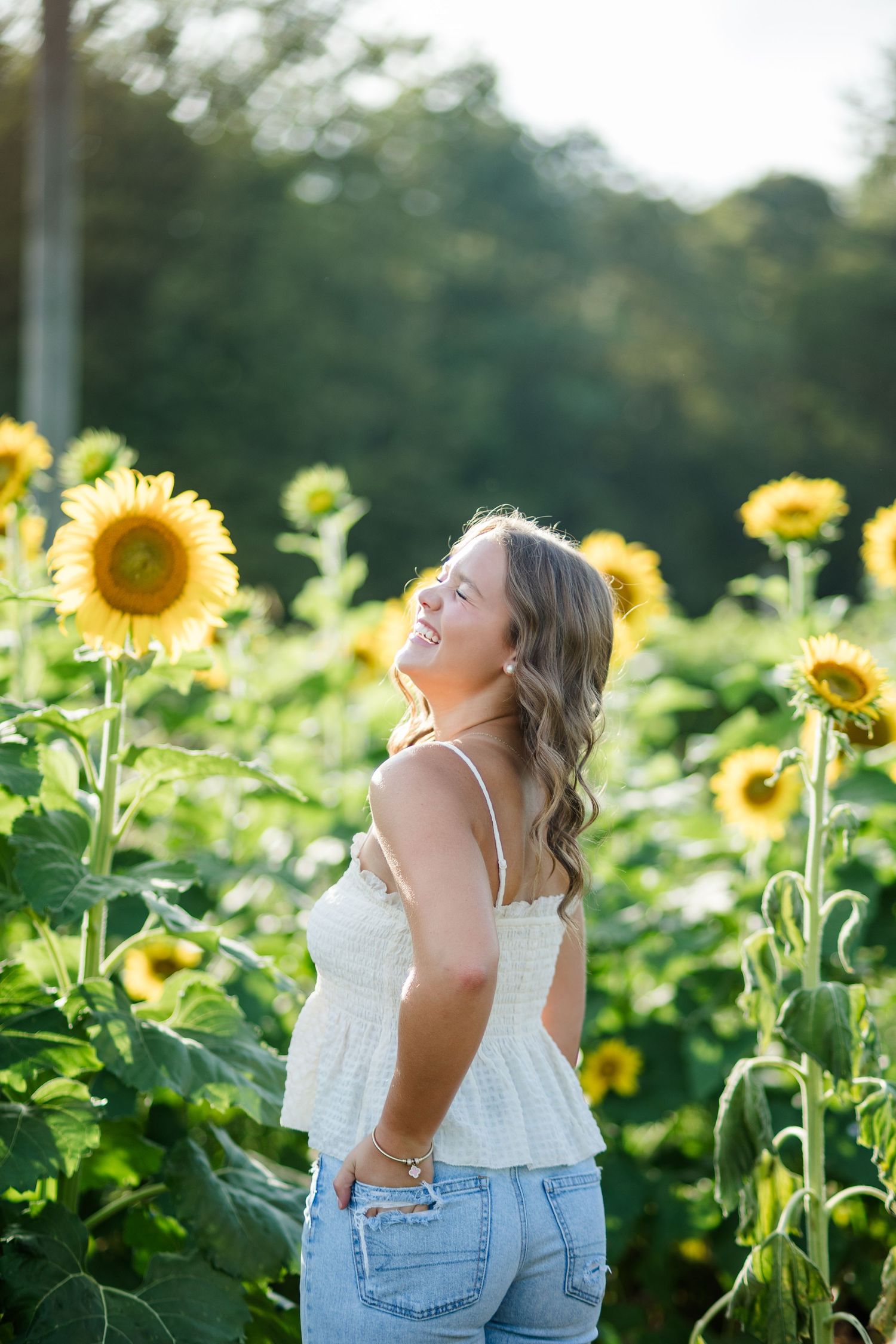 Young woman laughing joyfully in a sunflower field, wearing white top and jeans on a sunny day.