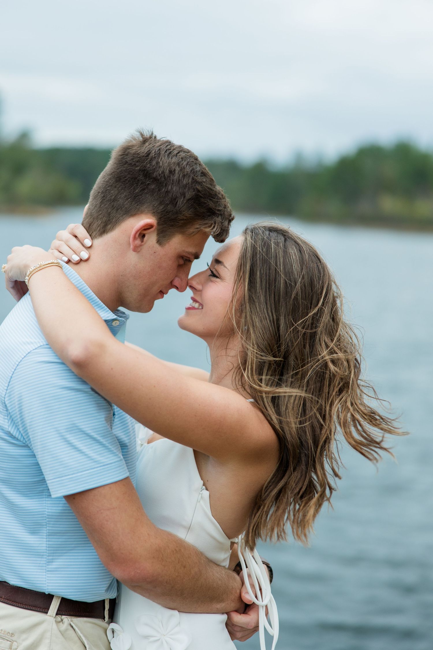 A smiling couple embraces and touches noses beside a calm lake, with a tree-lined shoreline behind them on a cloudy day.