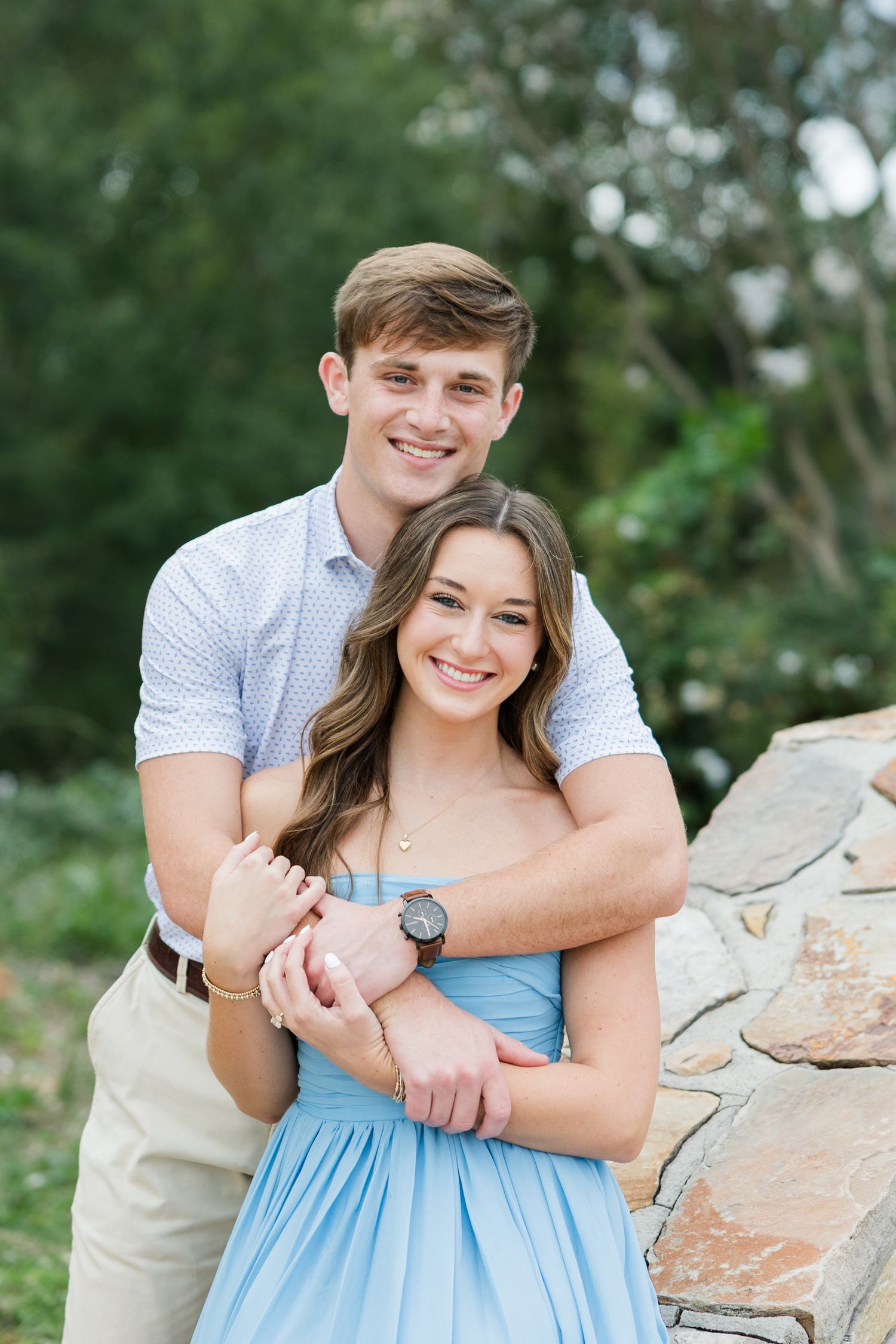 Smiling young couple posing outdoors as a man in a patterned shirt hugs a woman in a light blue dress from behind near a stone wall.