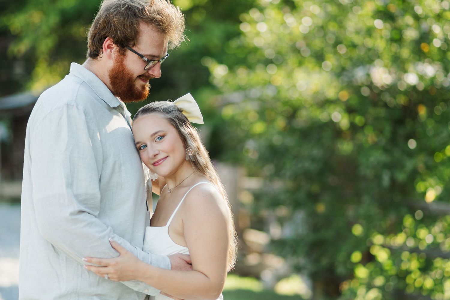 A couple embraces outdoors, woman in white dress with bow, man in light blue shirt, surrounded by lush green trees.