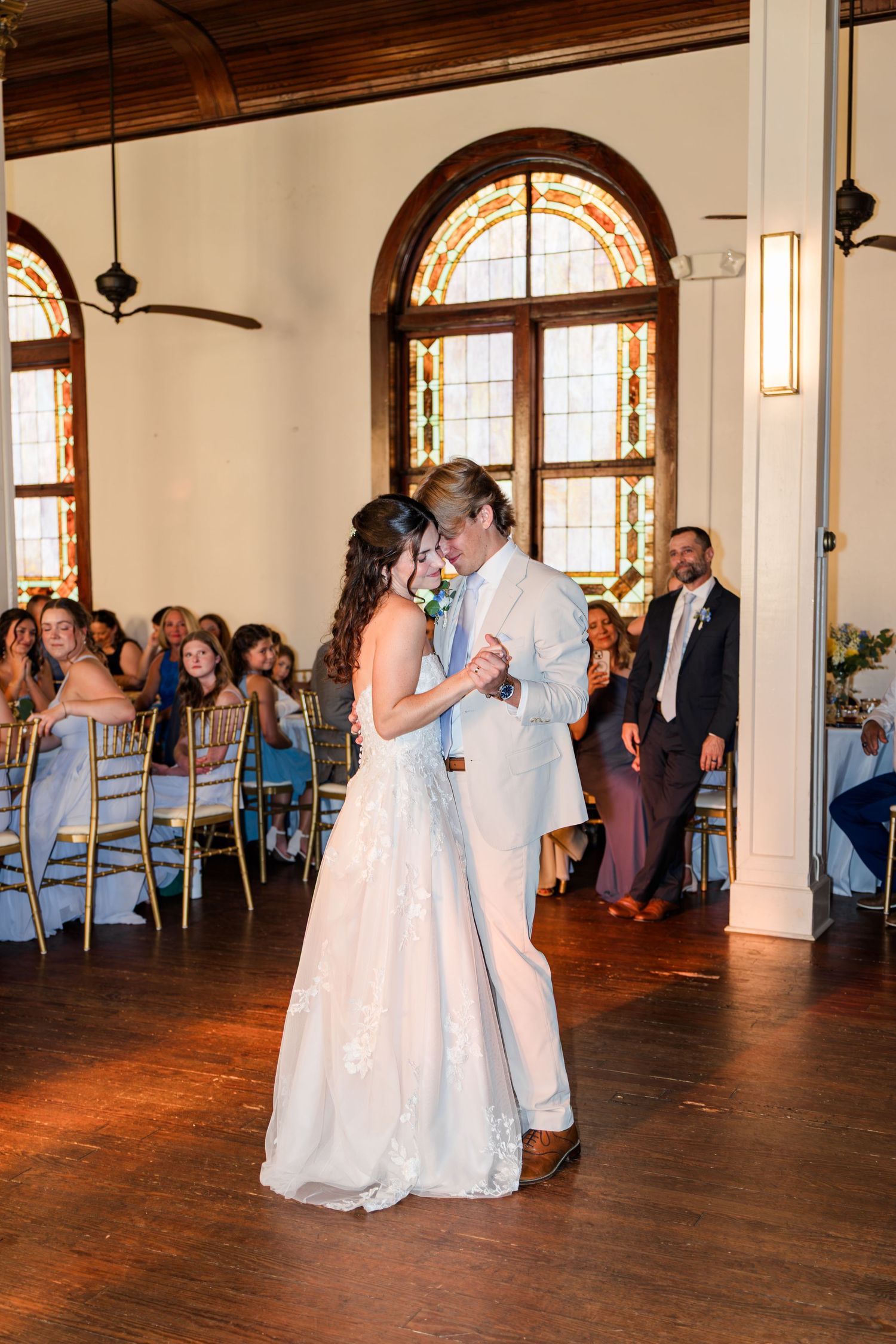 A bride and groom share their first dance in an elegant wedding venue with stained glass arched windows.
