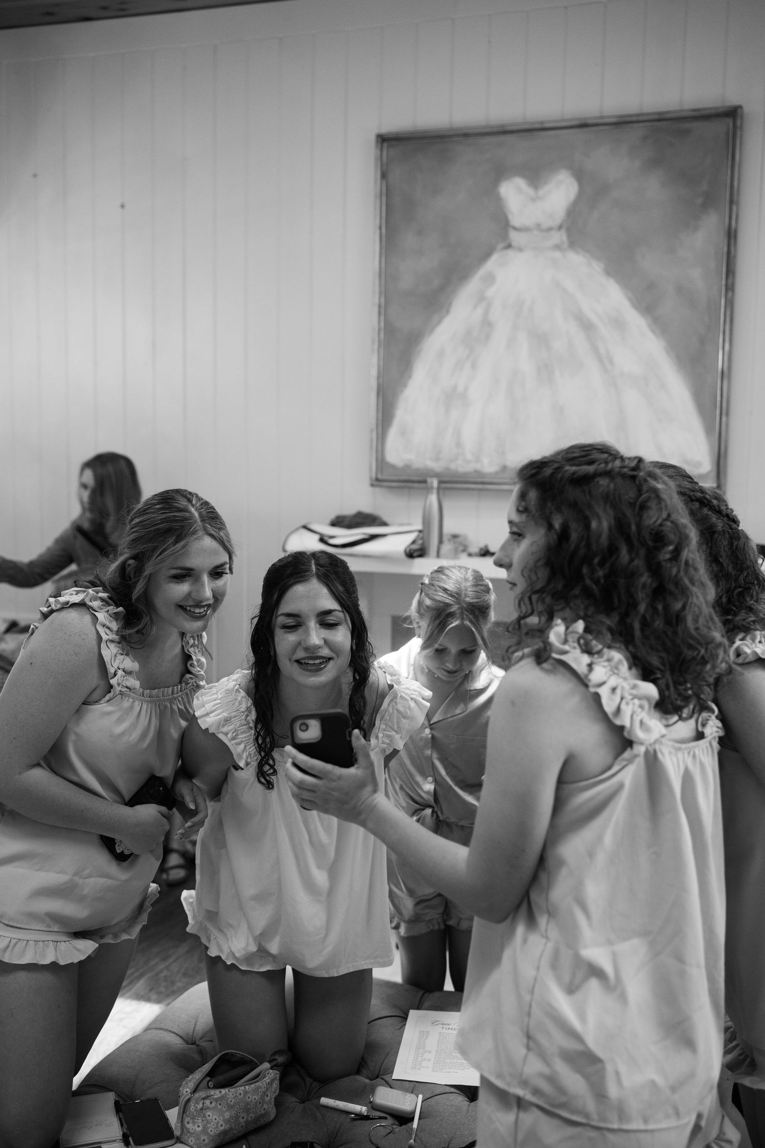 Bridesmaids in white pajamas laugh together looking at a phone during wedding preparations in black and white.