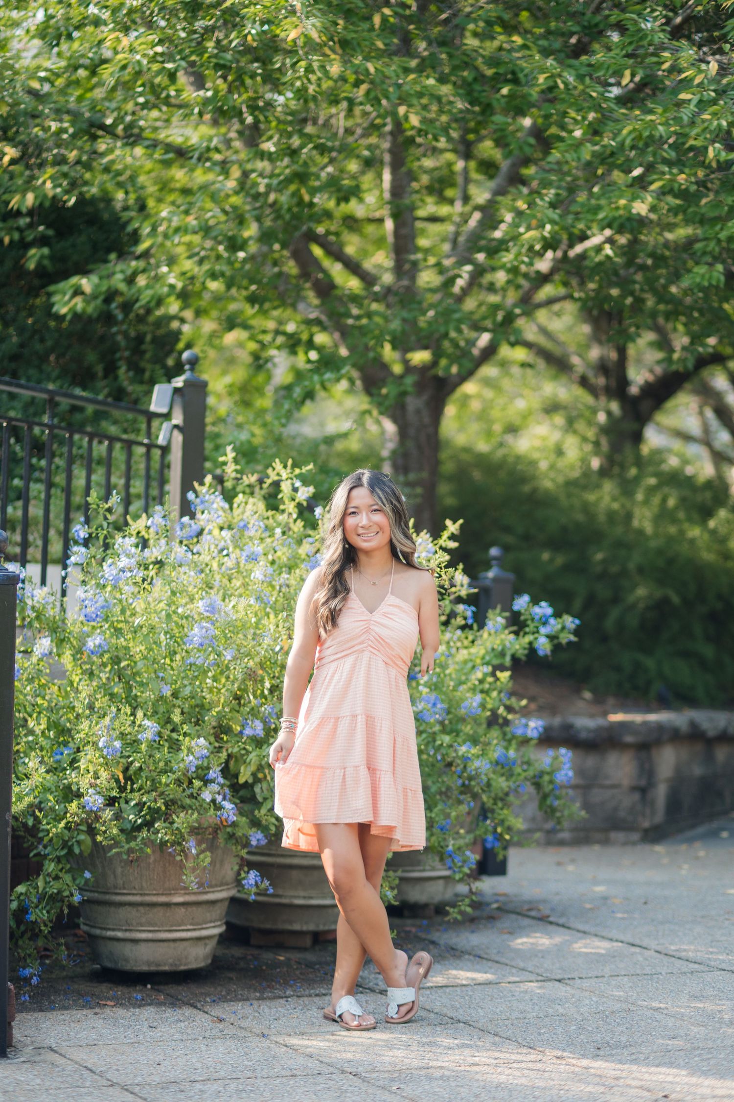 Young woman in peach sundress standing beside blue flowering potted plants outdoors.