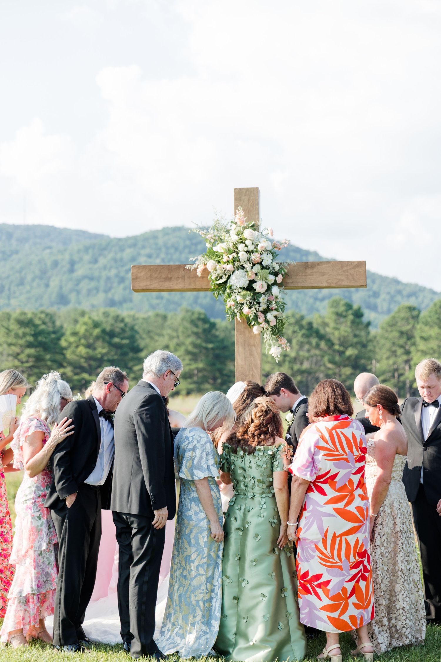 Wedding guests bow in prayer before a floral-adorned wooden cross at an outdoor ceremony with mountain views.