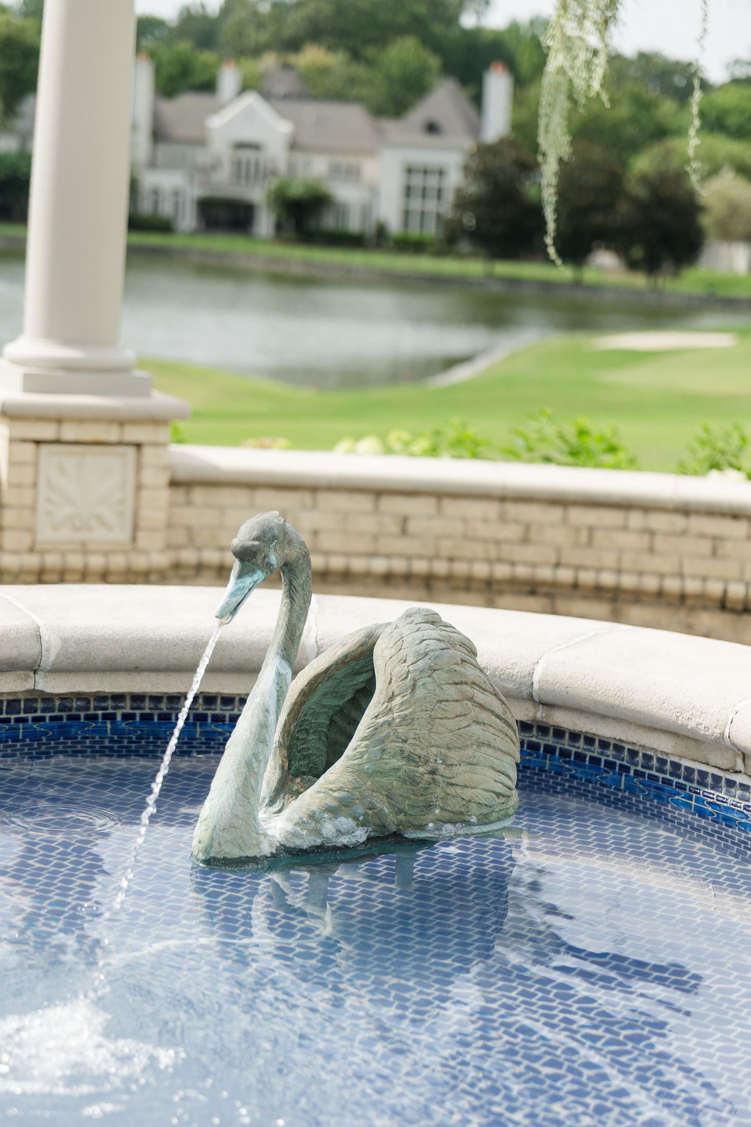 Bronze swan fountain sculpture spouting water in a blue mosaic pool with green golf course and lake in background.