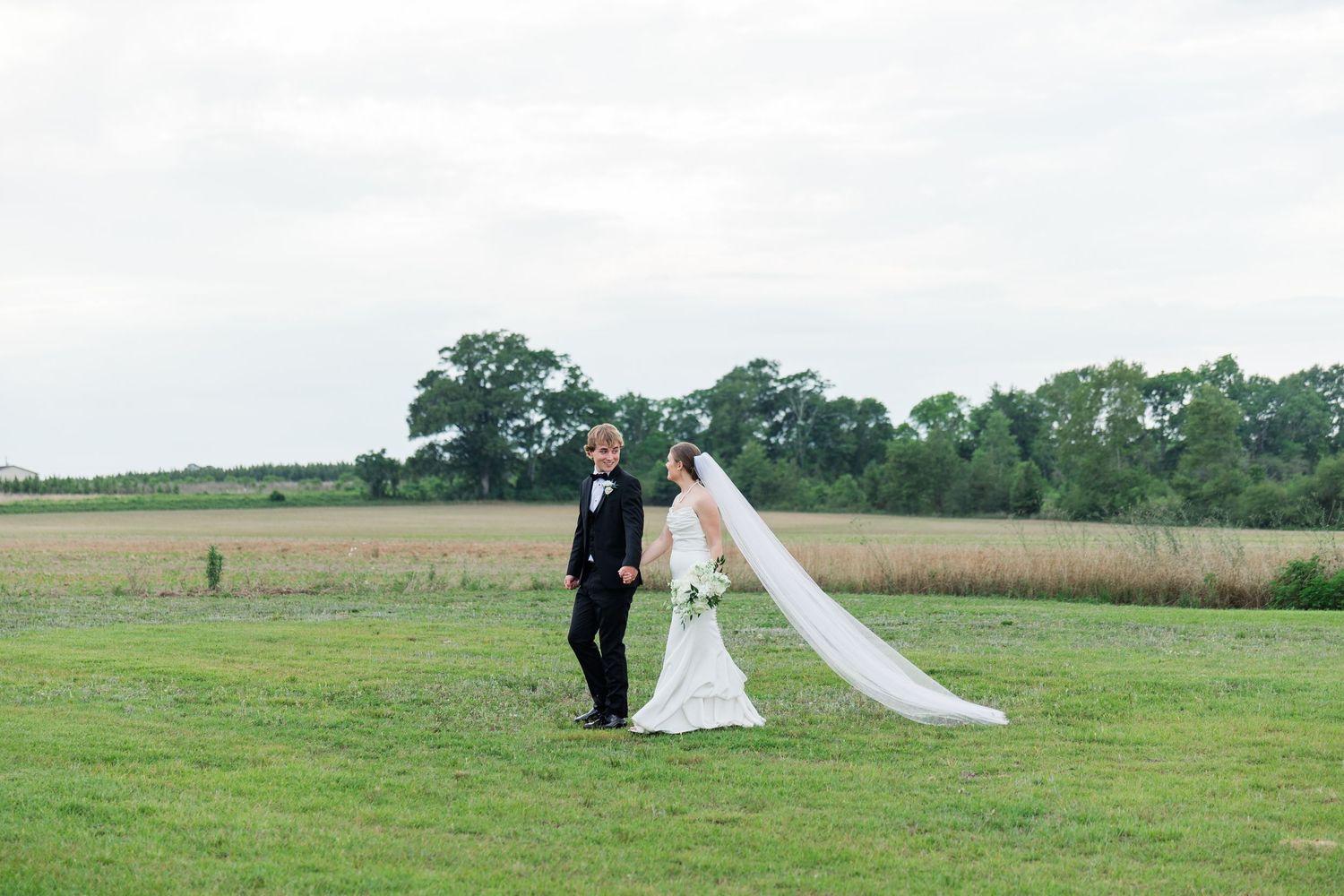 Bride and groom stand in open green field with long cathedral veil flowing in the wind.