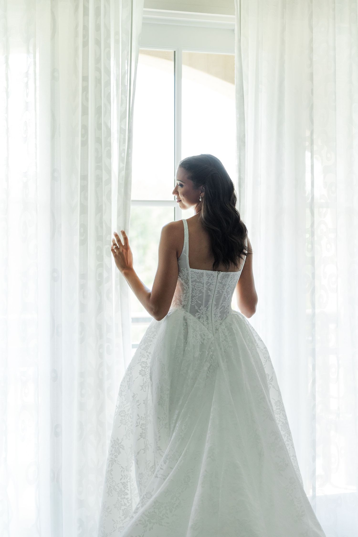 A bride in a white lace ballgown stands at a sunlit window, her back to camera, dark hair cascading down.