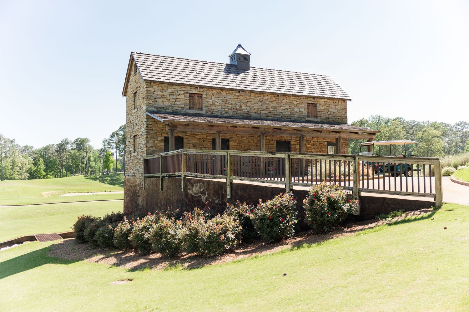 Historic two-story stone building with wooden deck surrounded by manicured green lawn and shrubs under clear blue sky.