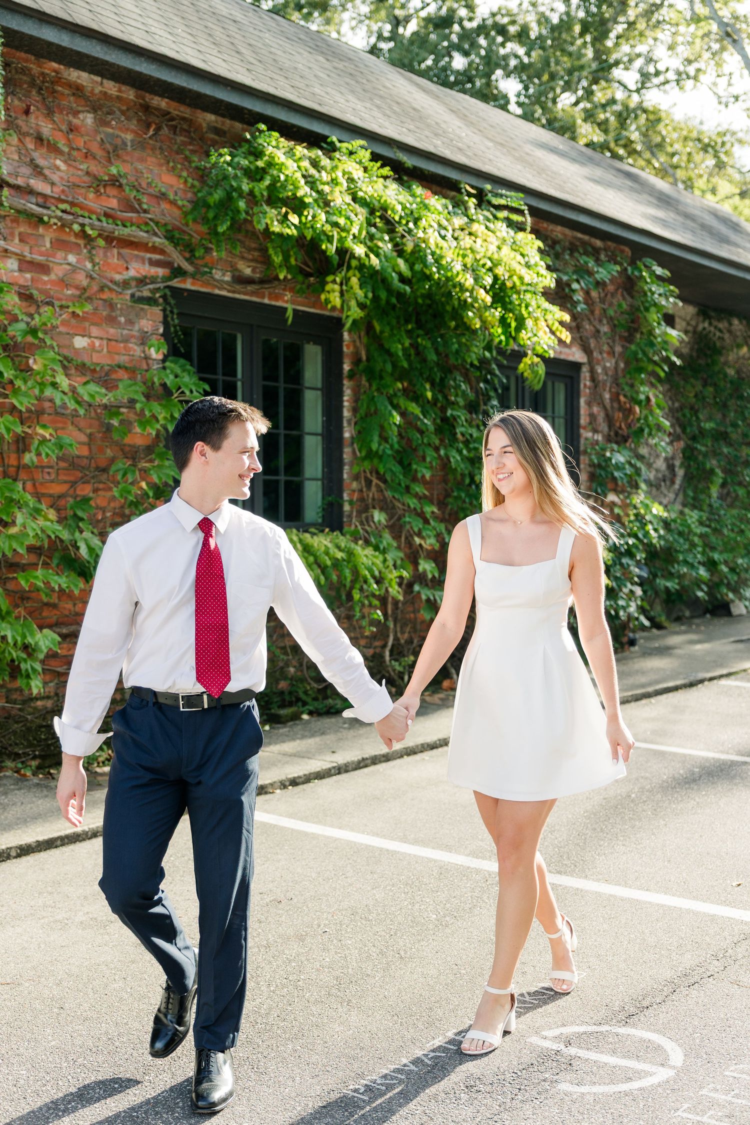 Couple holding hands walking past ivy-covered brick building on sunny day, man in white shirt red tie, woman in white dress.