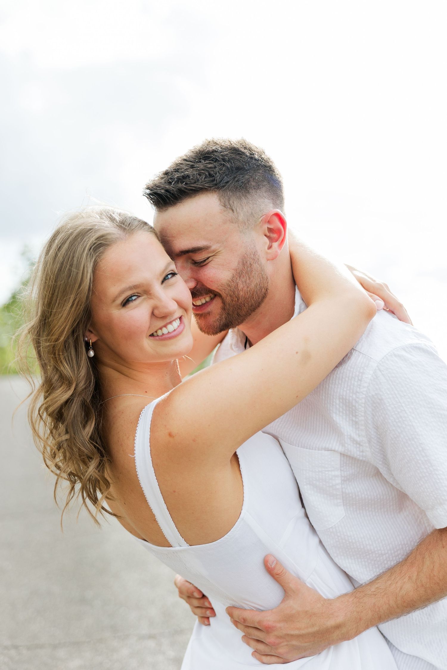 Smiling woman in white dress embraces man outdoors during romantic engagement photo session.