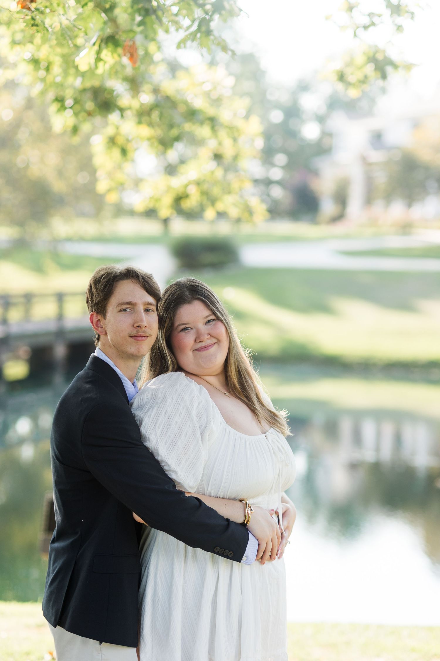 Couple embracing near a pond in a lush green park, man in navy blazer hugging woman in white dress.