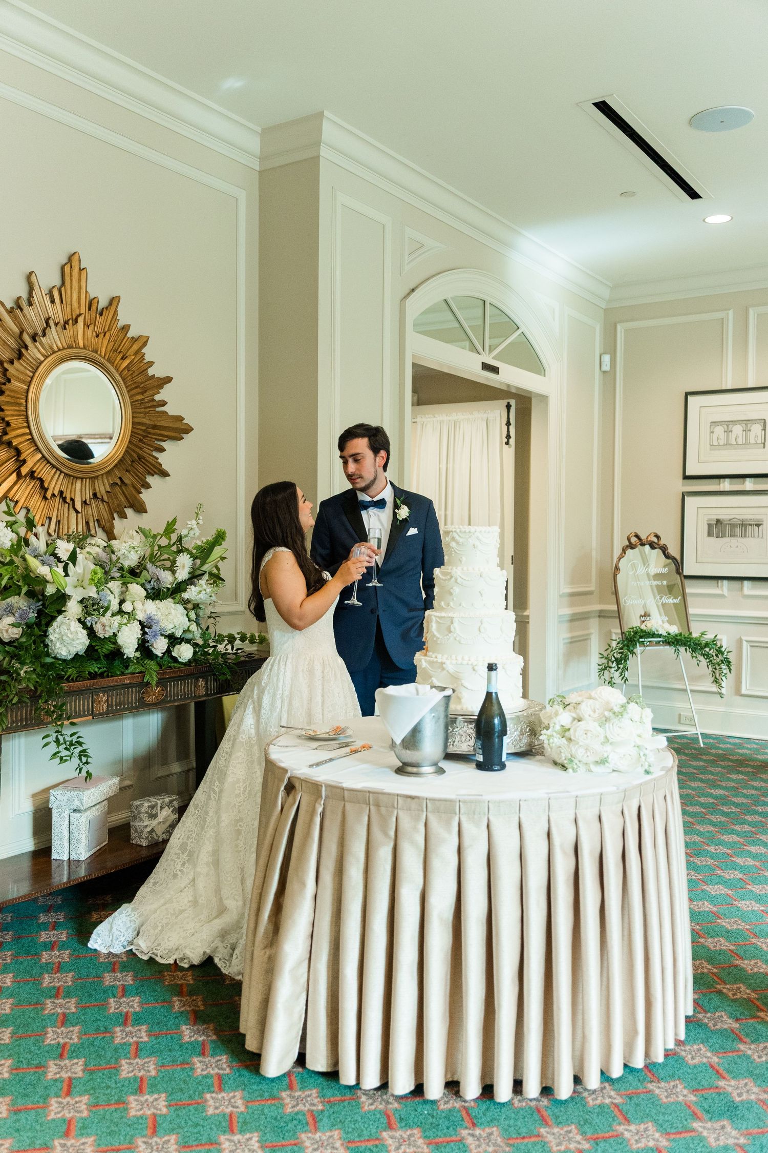 Bride and groom toasting champagne beside tiered white wedding cake in an elegant reception ballroom.