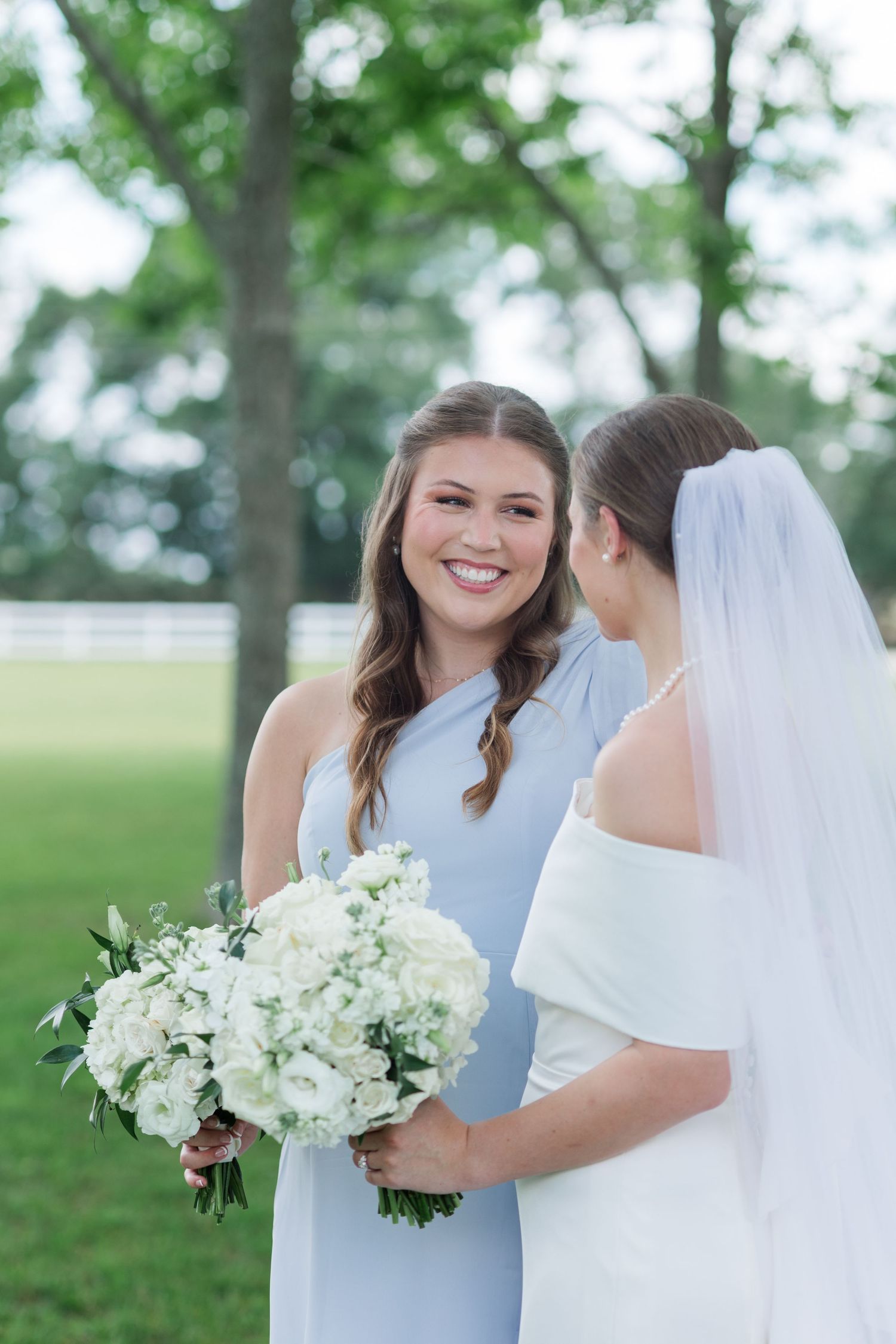 Smiling bridesmaid in light blue dress holds white floral bouquet beside bride in white gown and veil outdoors.