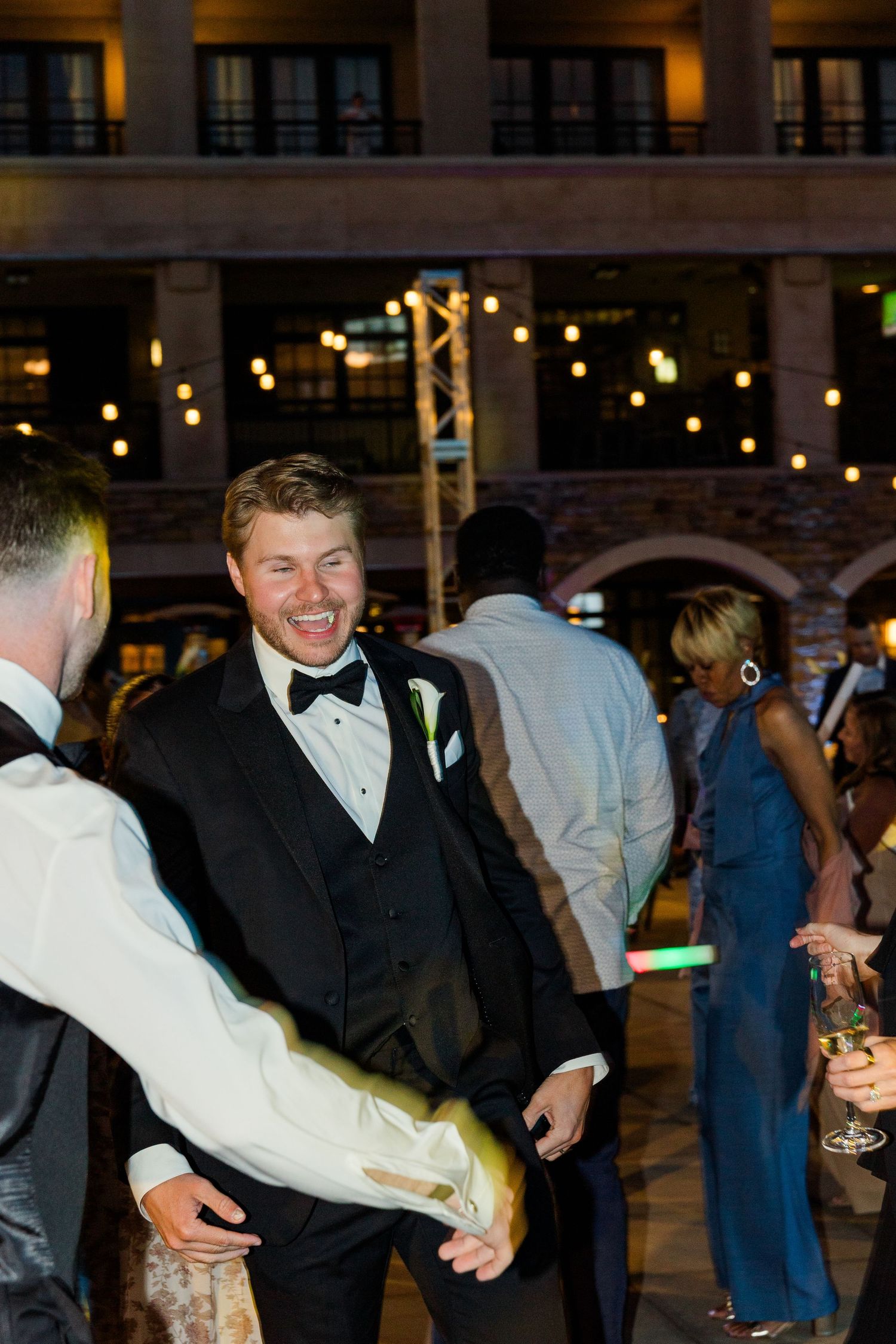Groom in black tuxedo with bow tie laughing and dancing at outdoor wedding reception with string lights.